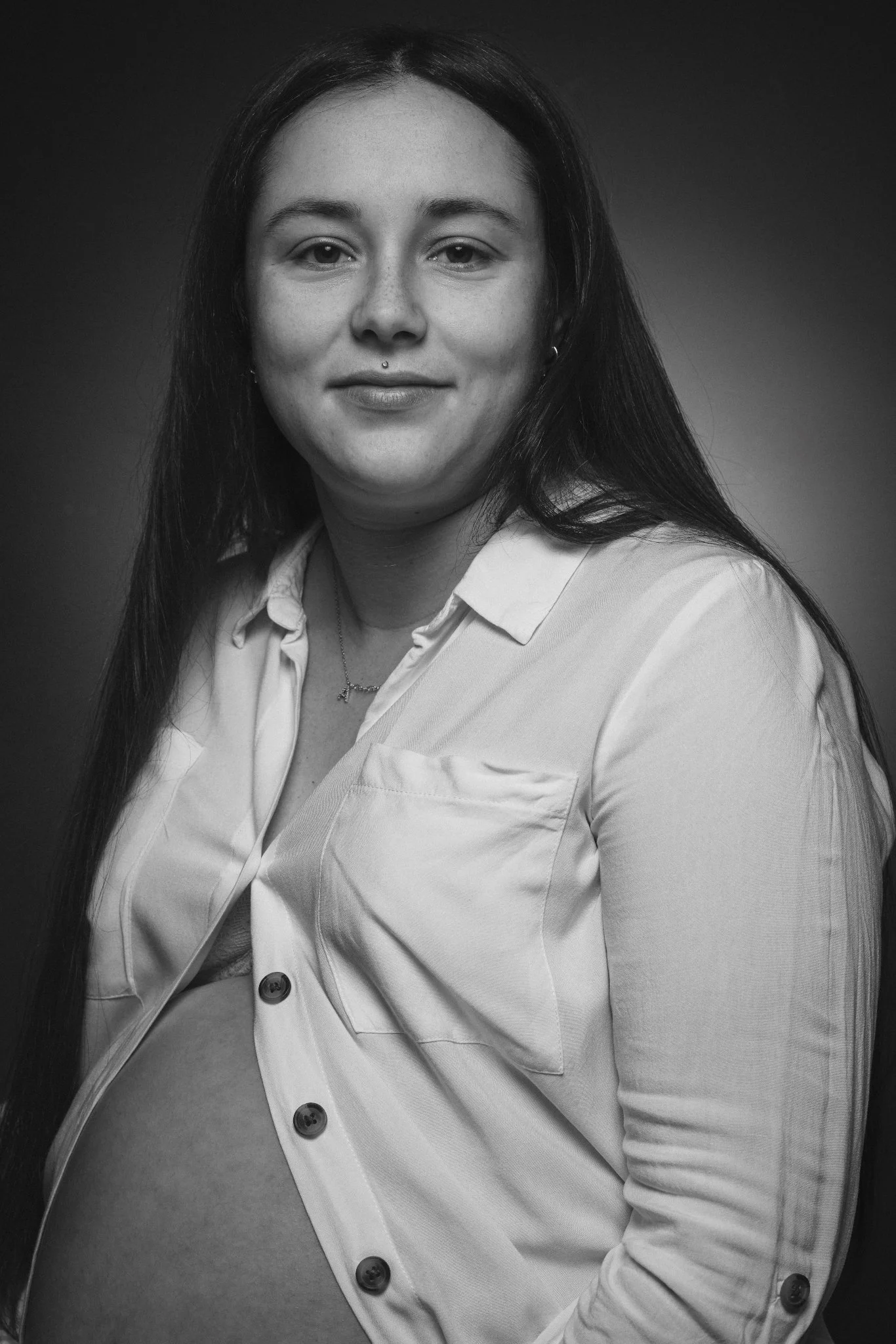 Schwarze und weiße Porträtaufnahme einer schwangeren Frau mit dunklen Haaren, weißen Hemd, die direkt in die Kamera schaut.