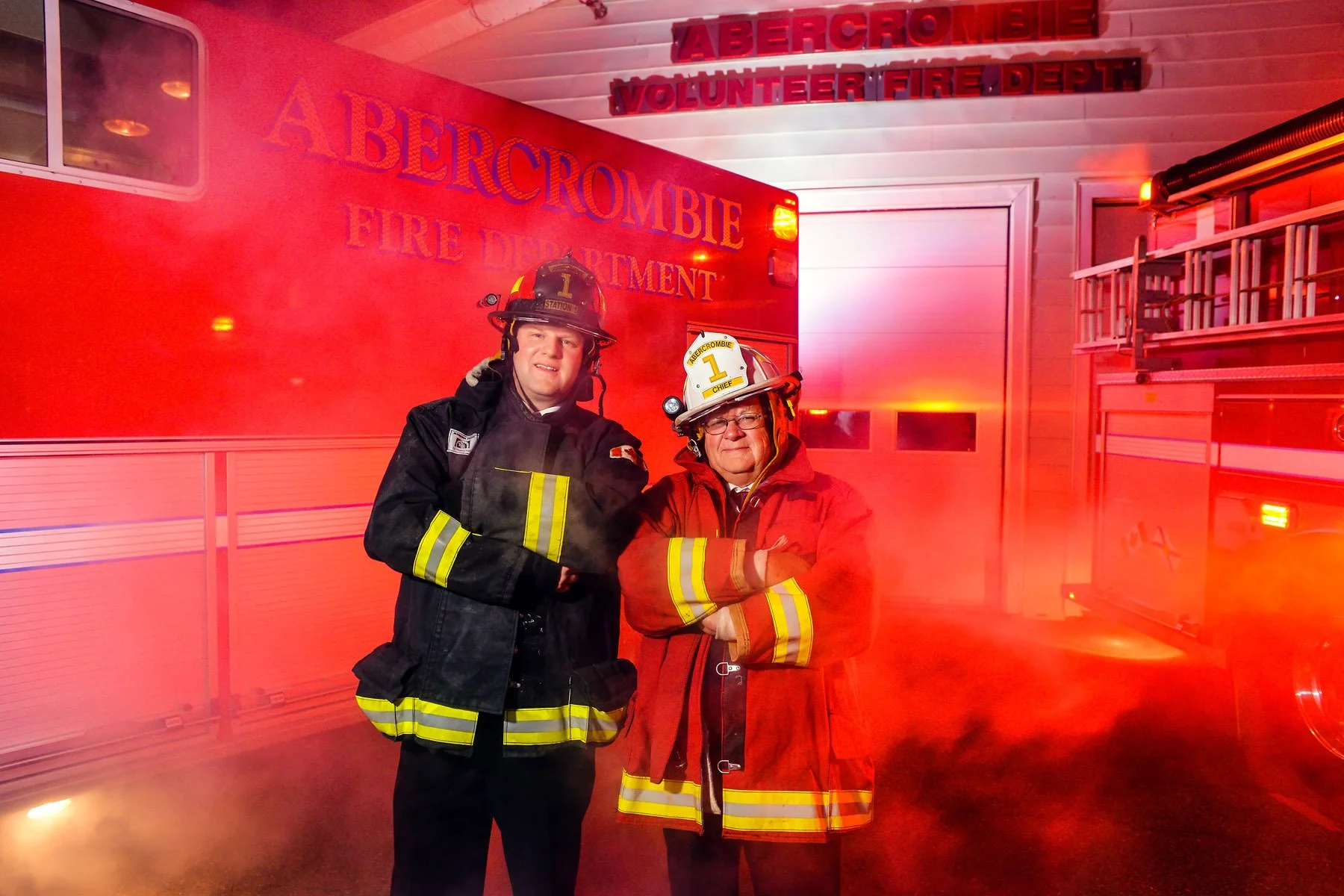 Two firefighters, one younger and one older, standing in front of a fire truck inside a fire station, wearing firefighting gear and helmets. The station has red emergency lights and signage reading "Abercrombie Volunteer Fire Department."