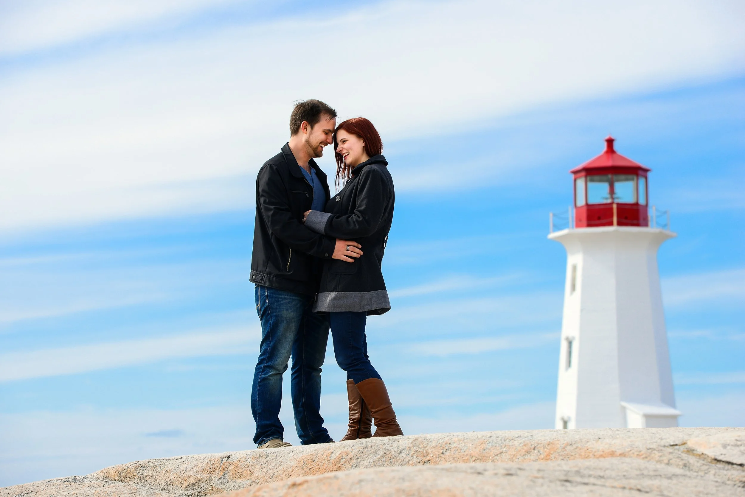 A couple smiling and standing close together on a rocky surface near a lighthouse against a blue sky.