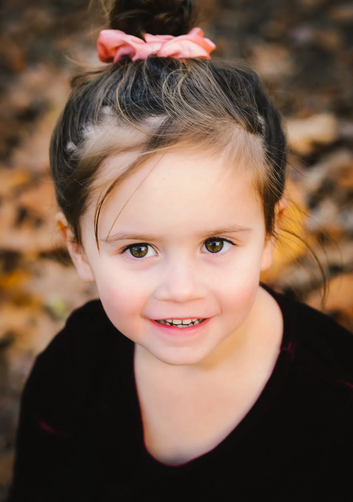 A young girl with brown hair tied in a bun with a pink hair scrunchie, smiling outdoors on a fall day with leaves in the background.