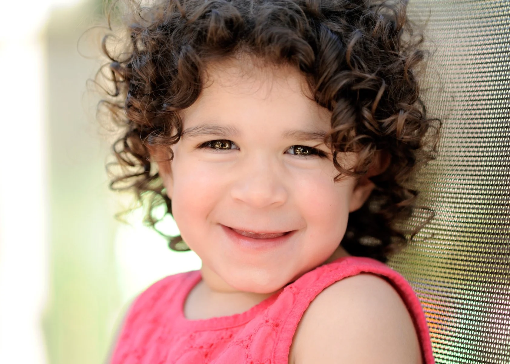 Close-up of a young girl with curly brown hair, smiling, wearing a pink sleeveless top, sitting outdoors against a blurred background.
