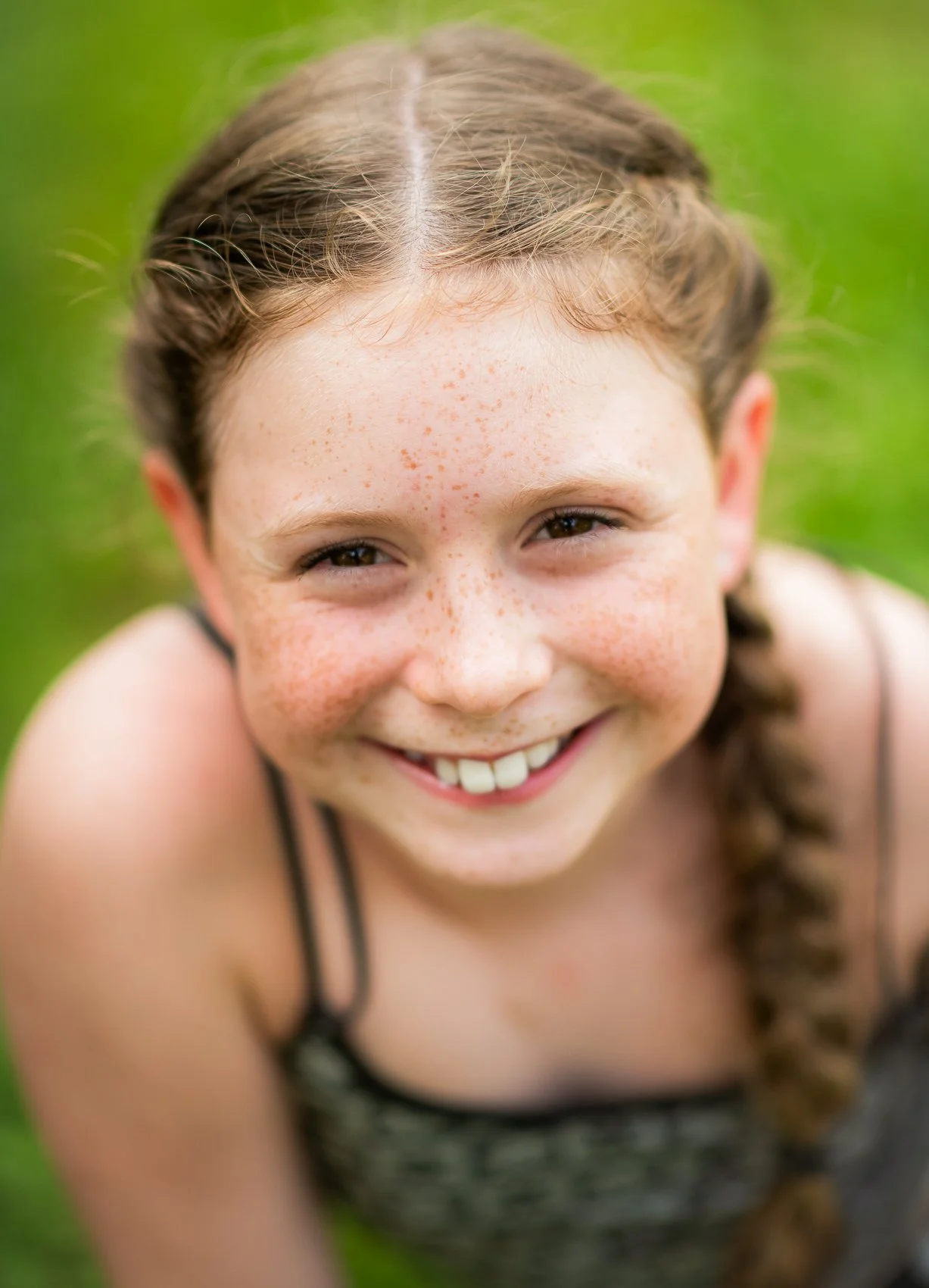 Close-up of a young girl with brown hair braided on the side and freckles, smiling outdoors with a green grassy background.