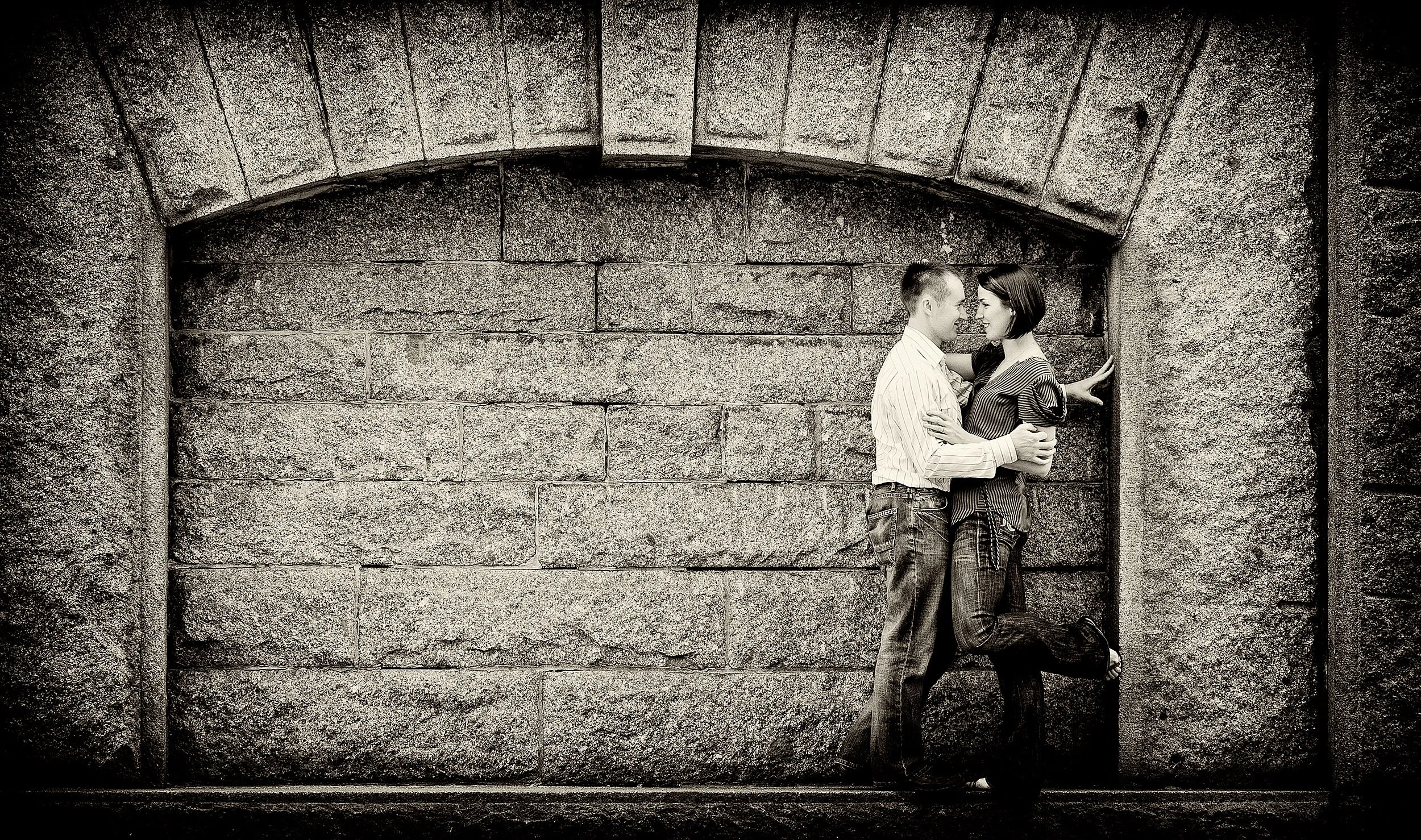 A black and white photo of a couple standing close and facing each other against a stone wall, touching foreheads and smiling.