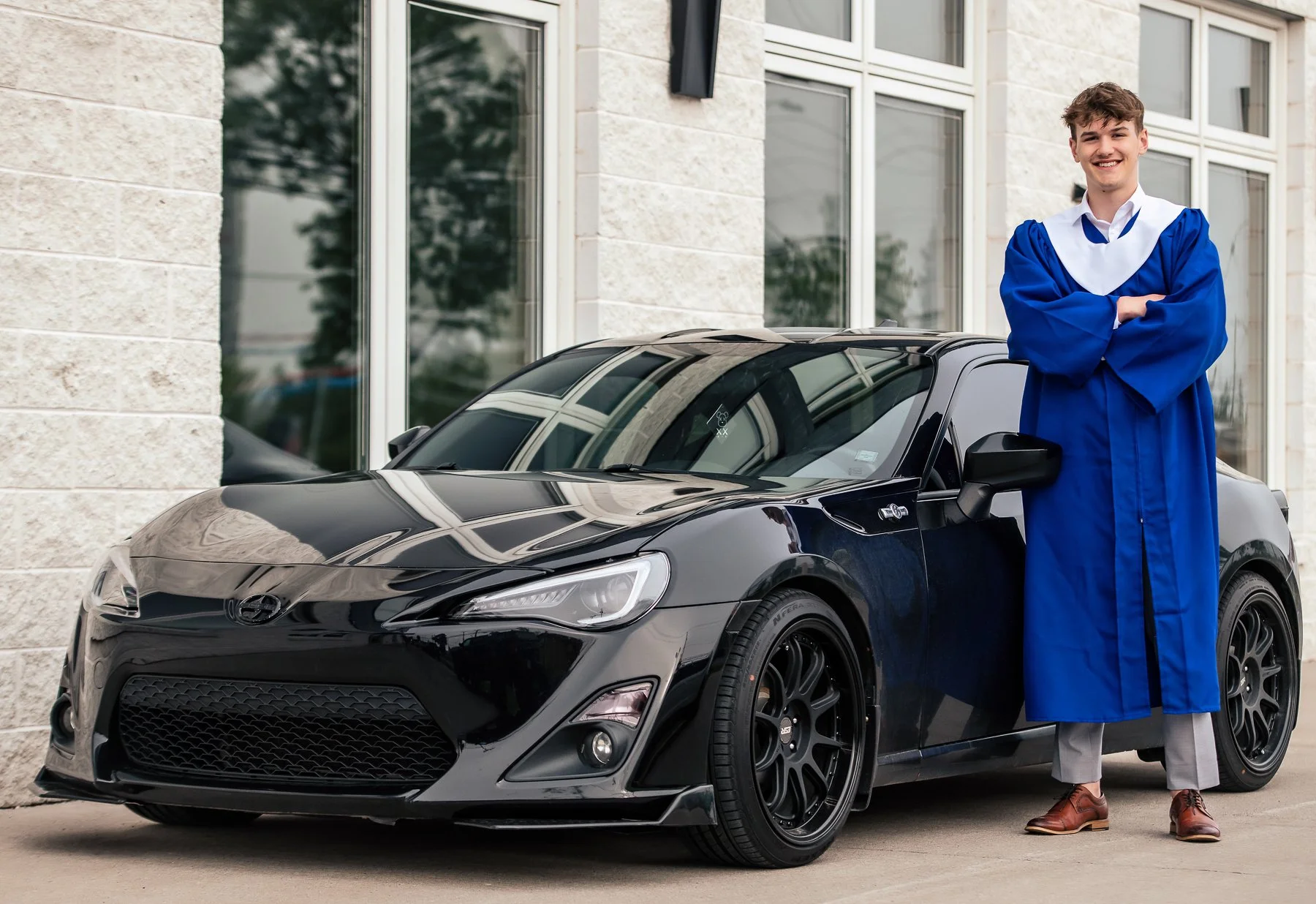 A young man in a blue graduation gown and hat standing with folded arms next to a black sports car in front of a modern building with large windows.