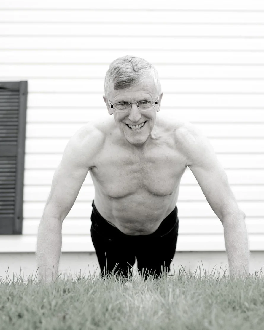 A smiling elderly man with glasses doing push-ups outdoors on grass in front of a house with horizontal siding.