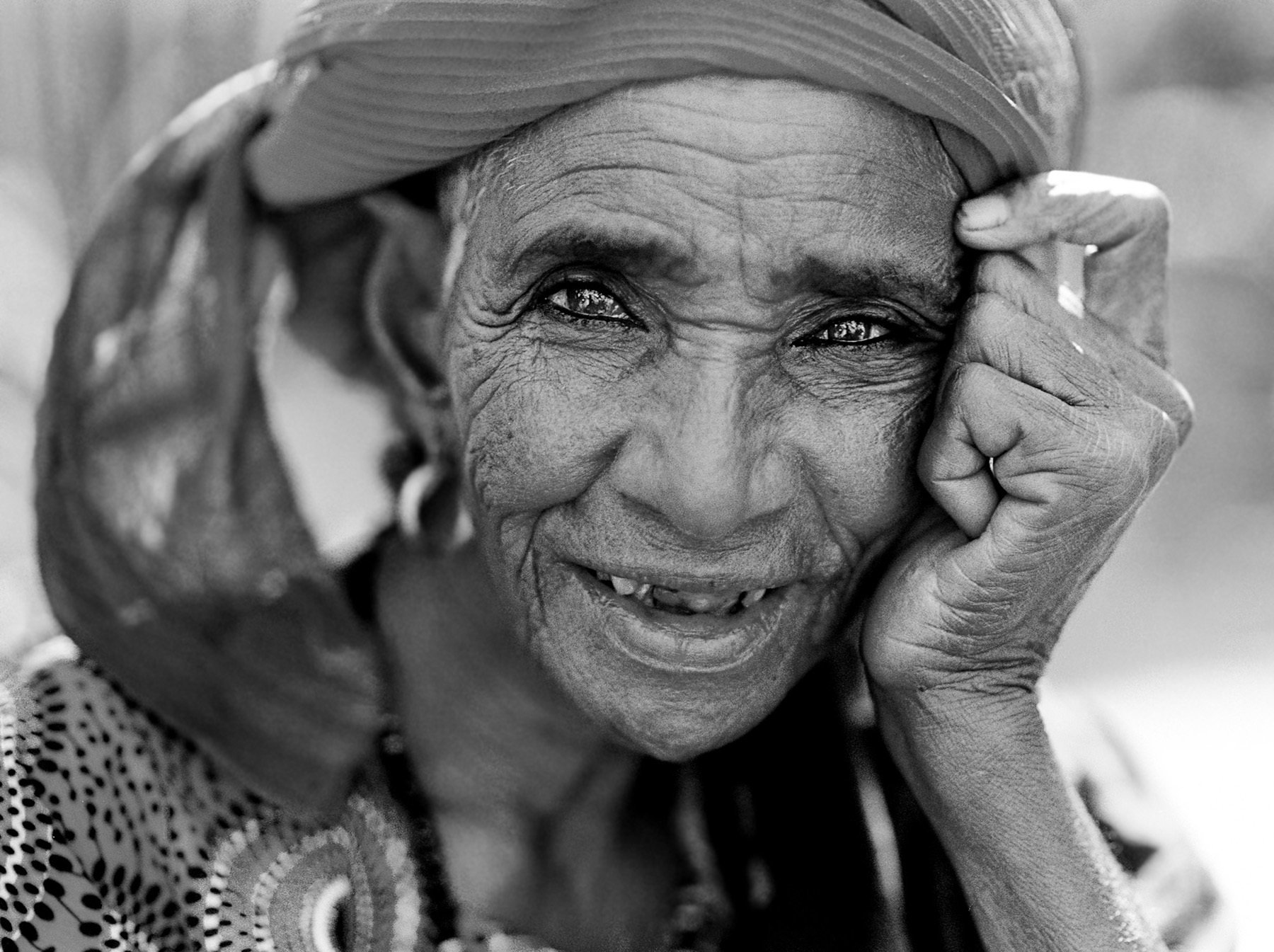 Close-up black and white photo of an elderly woman smiling, wearing a headscarf, with expressive eyes and visible wrinkles.