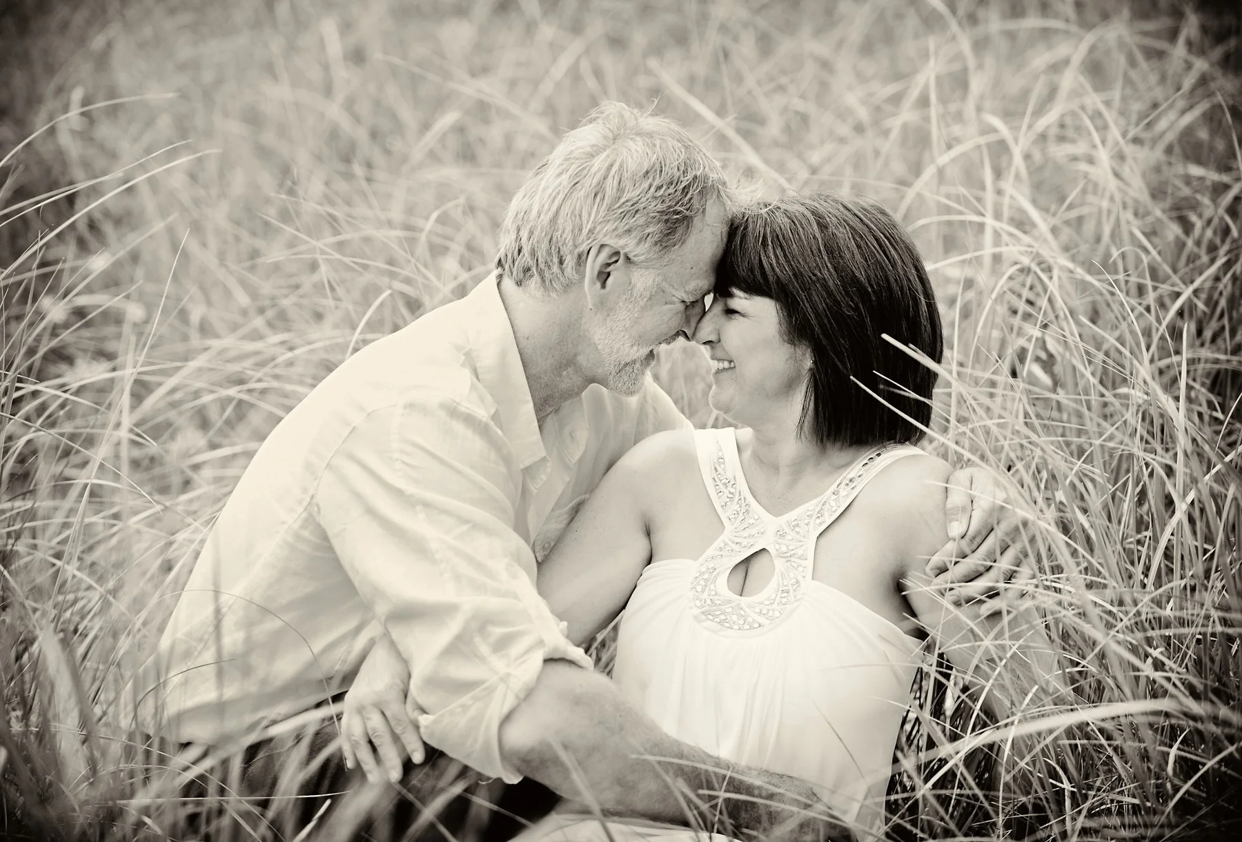 A black-and-white photo of a couple sitting in tall grass, touching foreheads and smiling affectionately, in an outdoor setting.