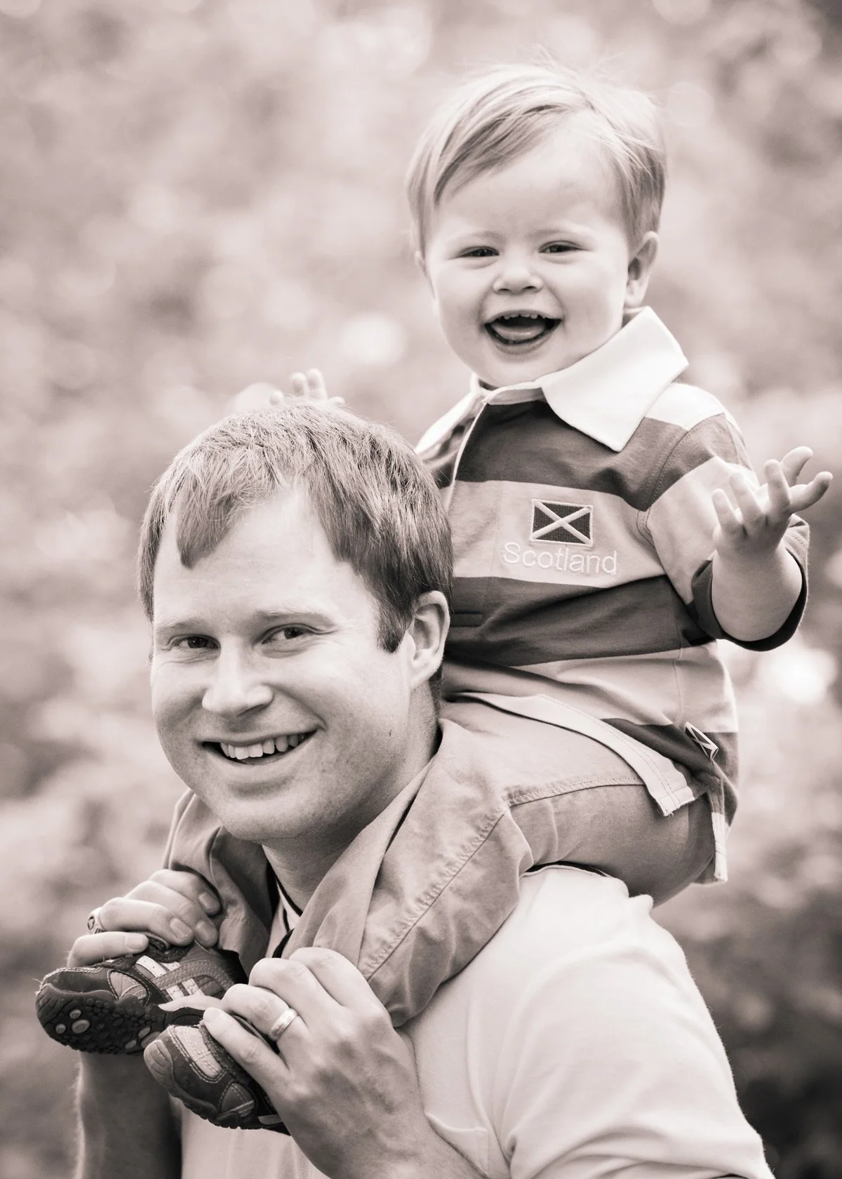 A smiling man with a young boy on his shoulders, outdoors with blurred trees in the background. The boy is wearing a jacket with a Scotland flag and the word 'Scotland' on it, and is waving his hand.