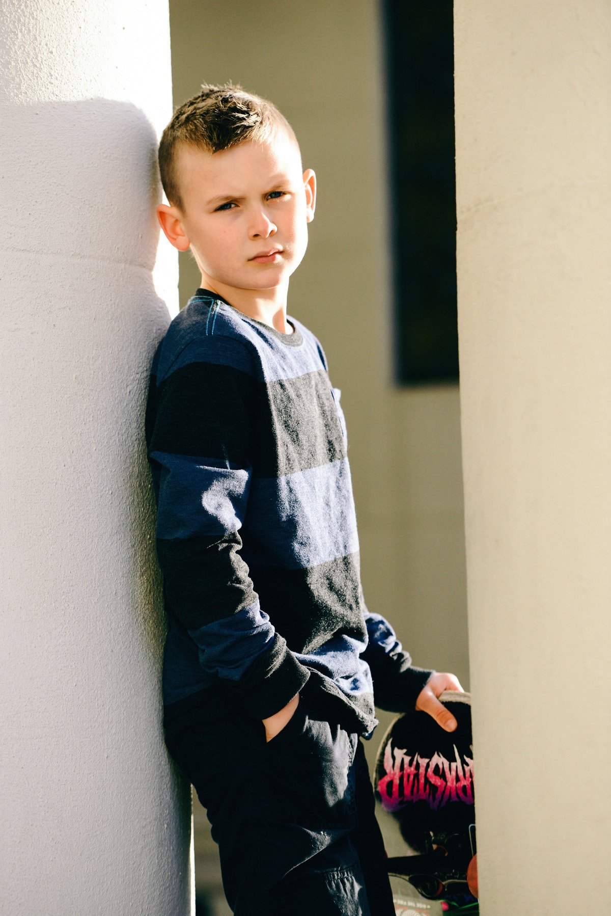 A young boy with short hair, dressed in a gray and black checkered long-sleeve shirt and dark pants, standing against a wall outdoors, holding a skateboard with pink and white graphics, and looking at the camera.