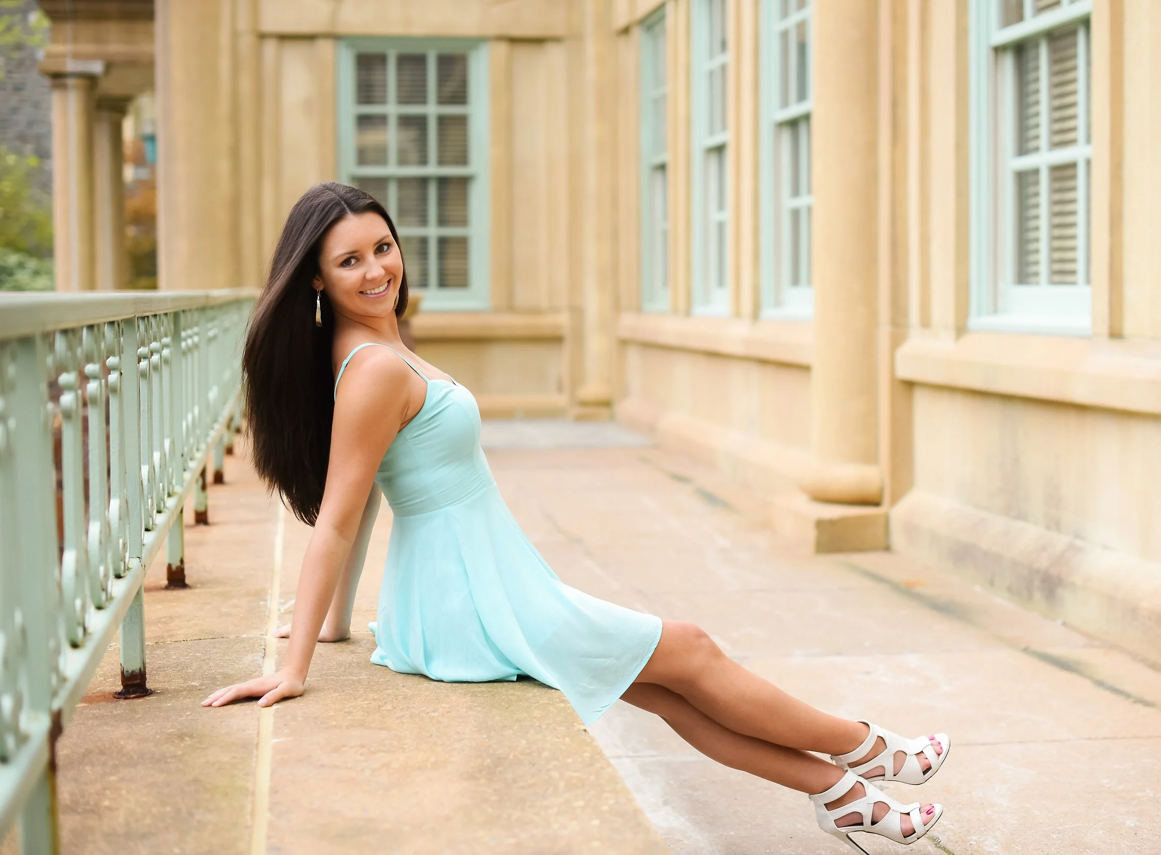 A young woman in a light blue dress and white high heels sitting on a stone ledge outdoors near a building with large windows.