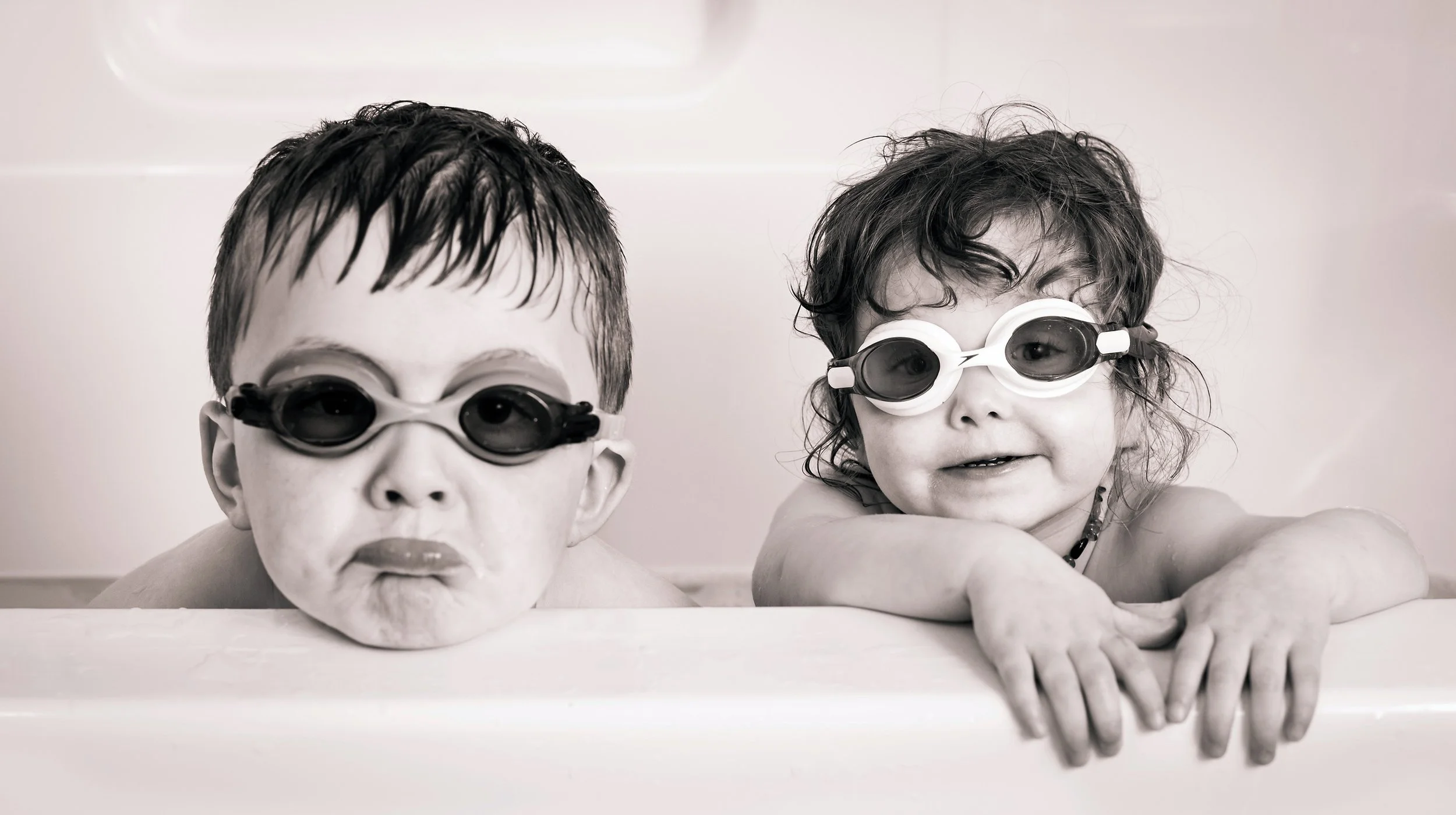 Two children wearing swimming goggles, leaning on the edge of a bathtub, smiling and looking at the camera.