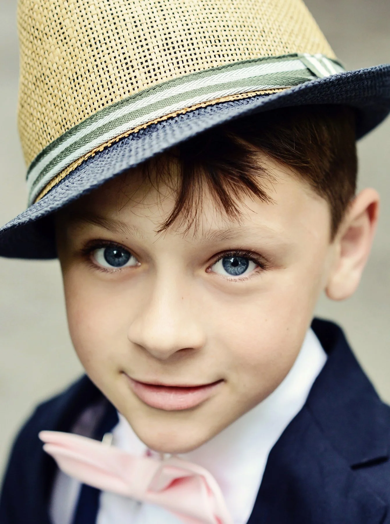 Close-up of a young boy with blue eyes, wearing a straw hat with a striped ribbon, a navy blazer, and a pink bow tie.