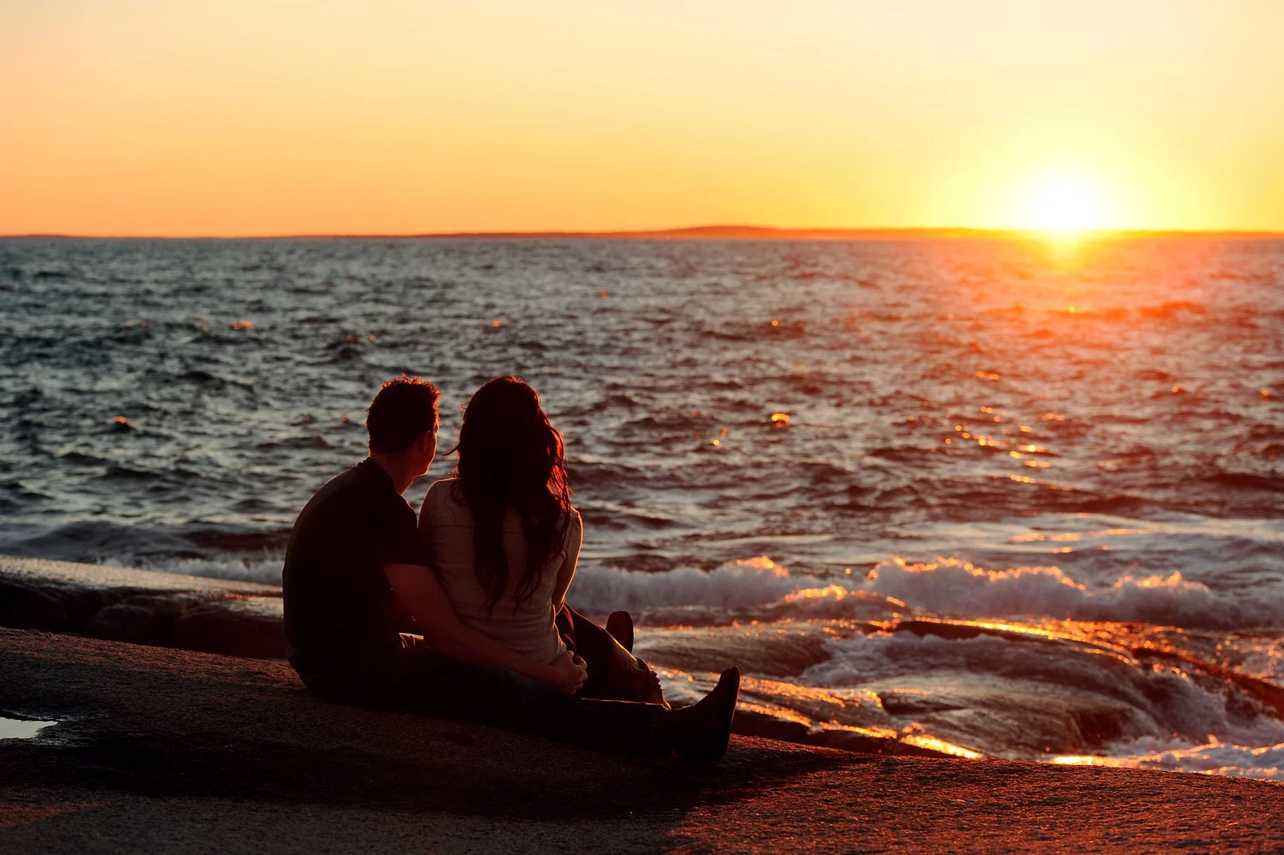 A couple sitting on a rocky beach at sunset, gazing at the ocean and the setting sun on the horizon.