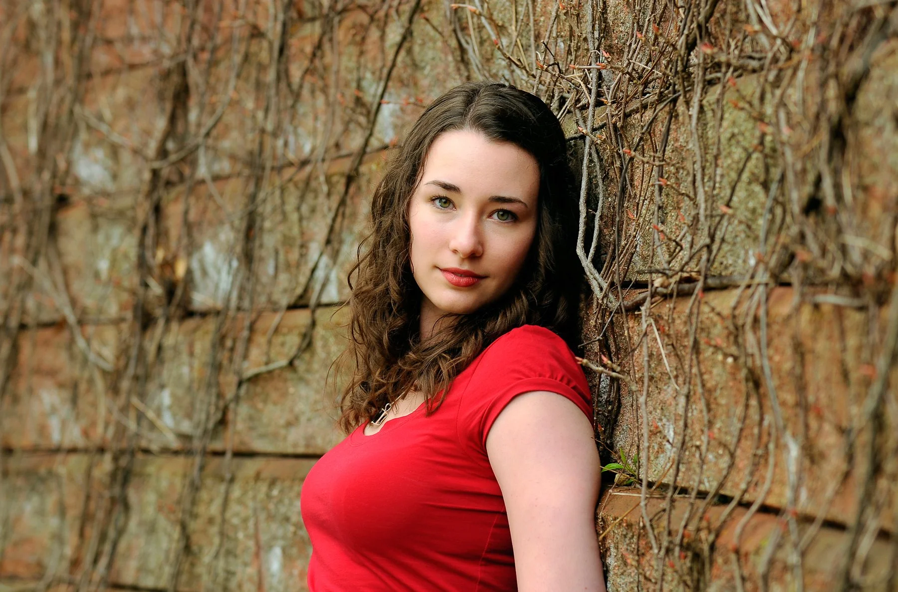 A woman with curly brown hair wearing a red top leaning against a wall covered with brownish, dried vines.