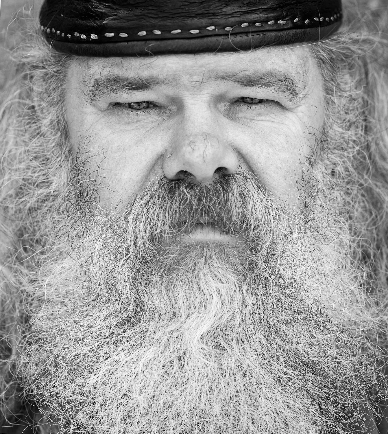 Close-up of an older man with a long, white beard, mustache, and curly hair wearing a hat, captured in black and white.