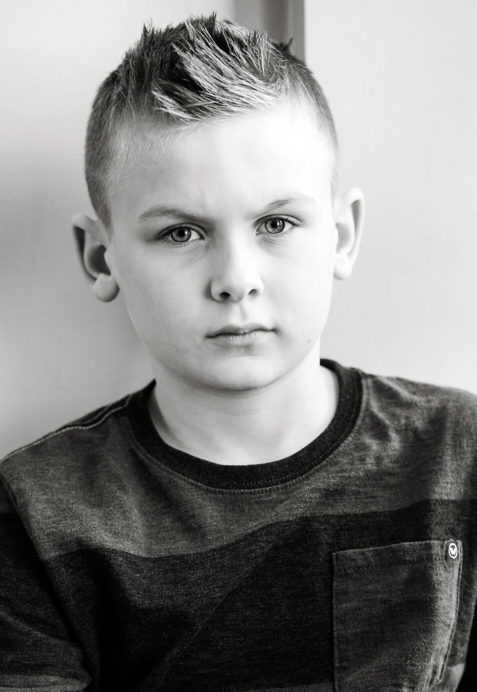 A young boy with short, spiked hair looking directly at the camera. He's wearing a dark t-shirt with a small logo on the chest. Black and white photo.