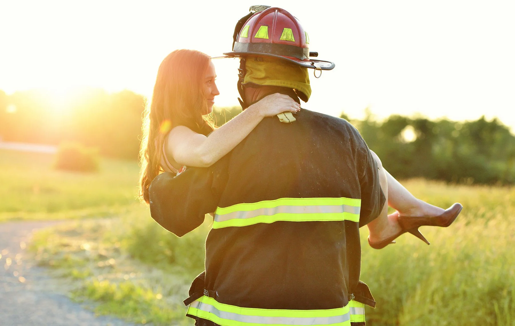 A firefighter carrying a woman in a field during sunset, with the woman holding onto the firefighter's shoulders.