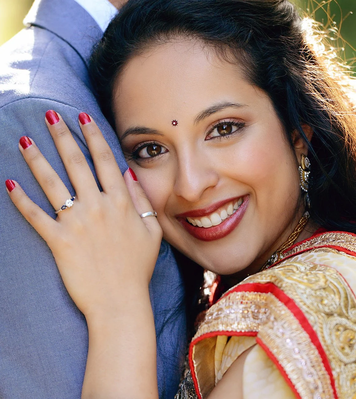 A smiling woman in traditional Indian attire hugging a person in a blue shirt. She has long black hair, brown eyes, and is wearing jewelry and red lipstick.