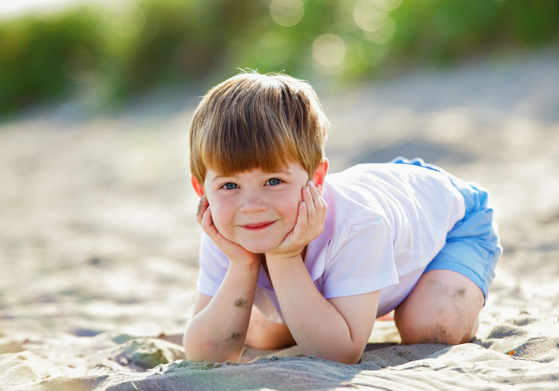 Young boy with brown hair and blue eyes lies on his stomach on the sandy beach, propping his head up with his hands, smiling at the camera, with some dirt on his arms and sandy background.