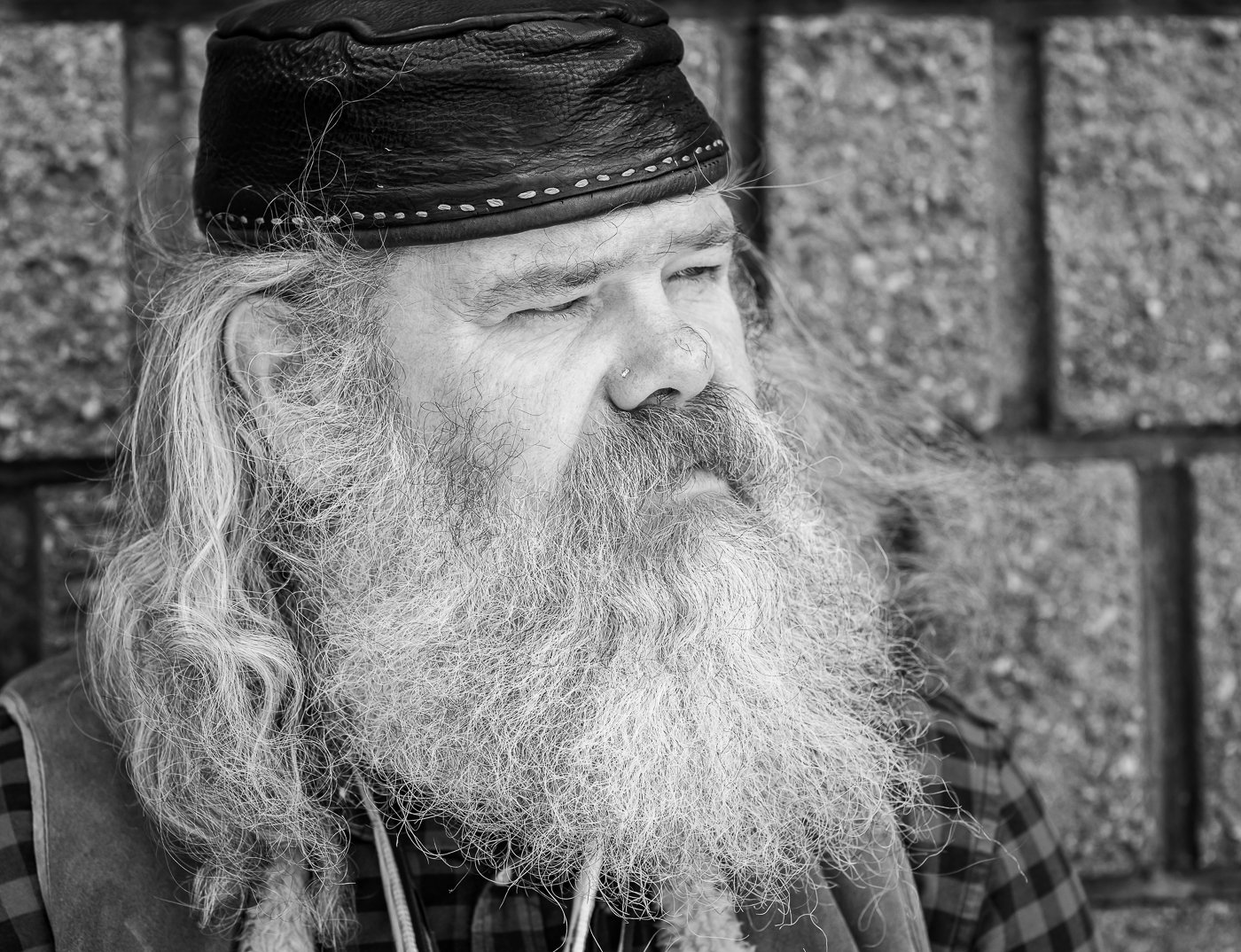 A black and white photo of an older man with long, gray hair and a full beard, wearing a black leather cap and looking to the side.