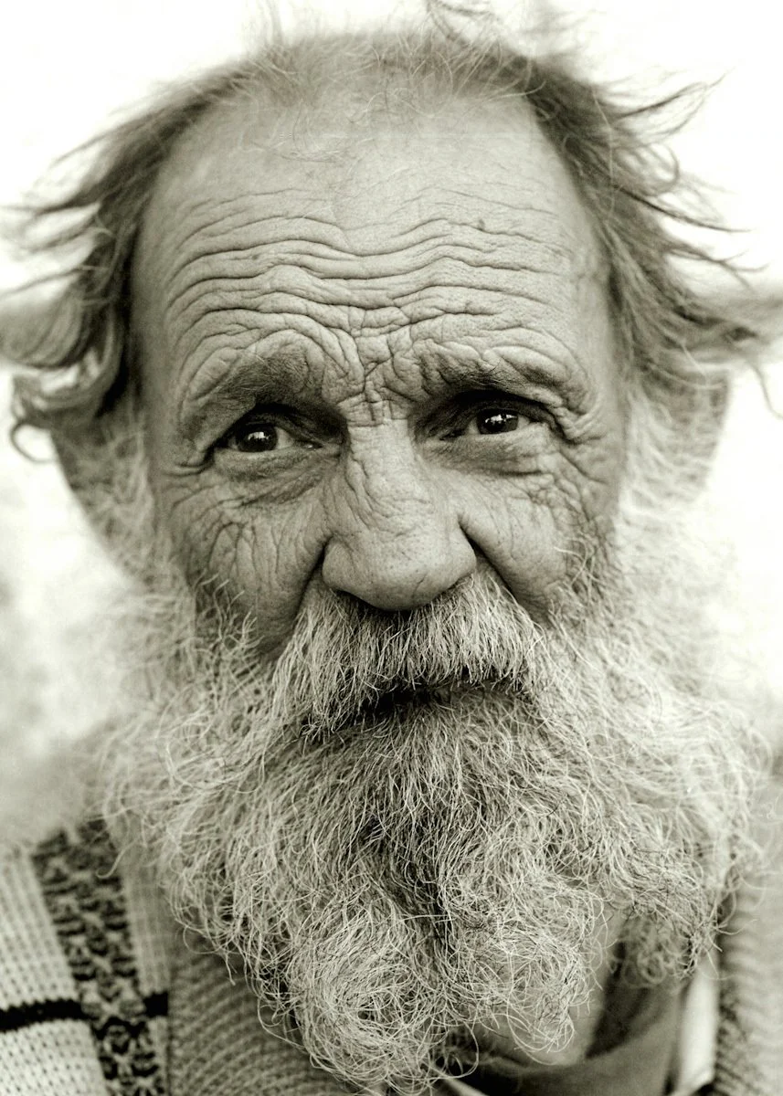 Close-up black-and-white portrait of an elderly man with a long, thick beard, wrinkled skin, and wild, unkempt hair, showing a gentle expression.