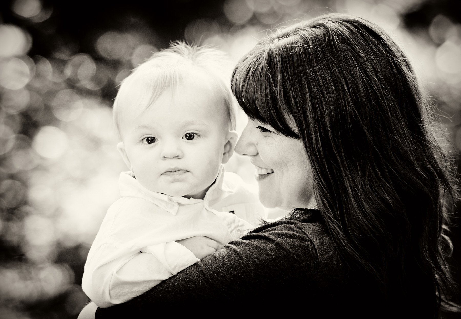 A woman joyfully holds a young child close, both smiling, with a blurred background of bokeh lights in black and white.