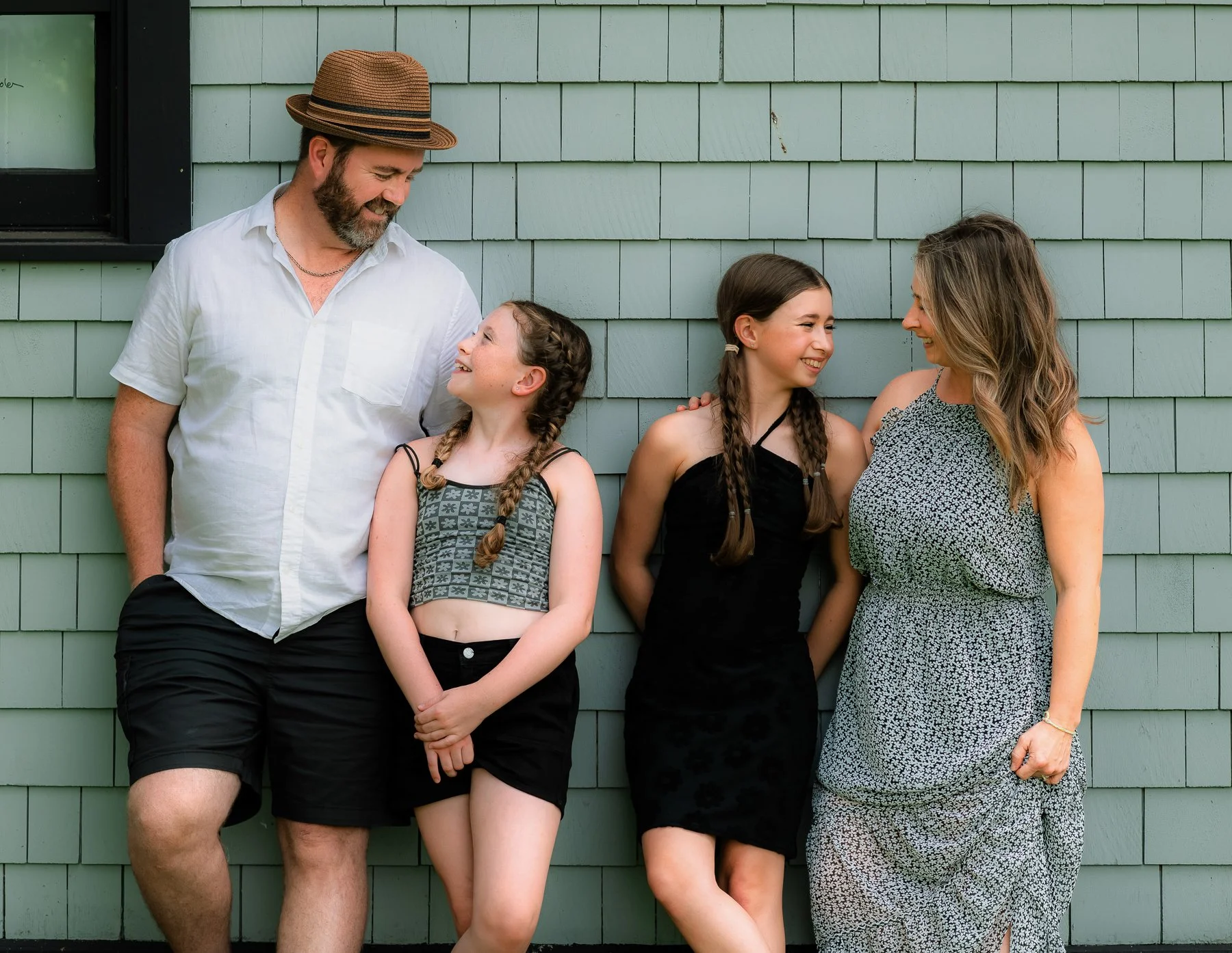 Family of four standing against a green shingled wall, smiling and looking at each other.