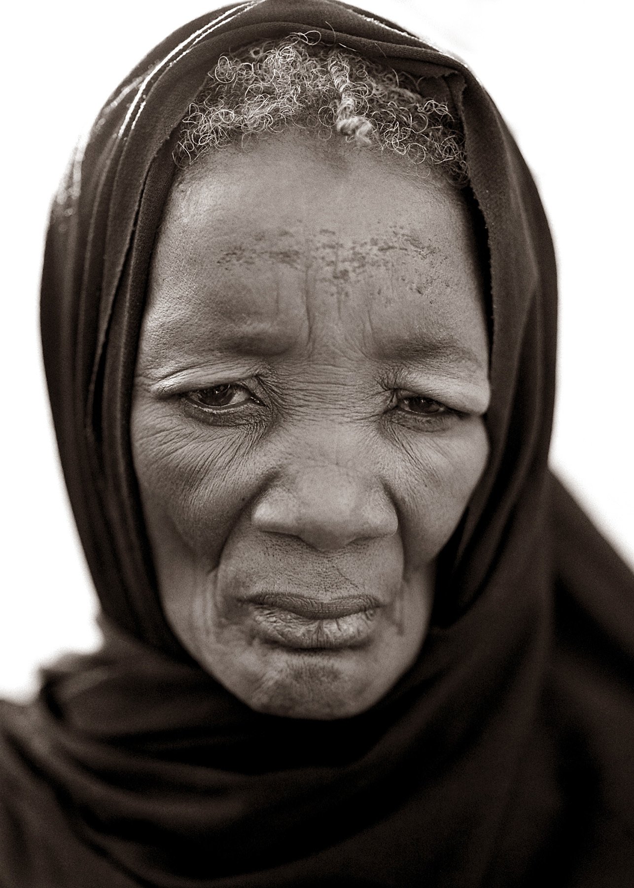 Black and white close-up portrait of an elderly woman wearing a headscarf, with visible facial wrinkles and a serious expression.