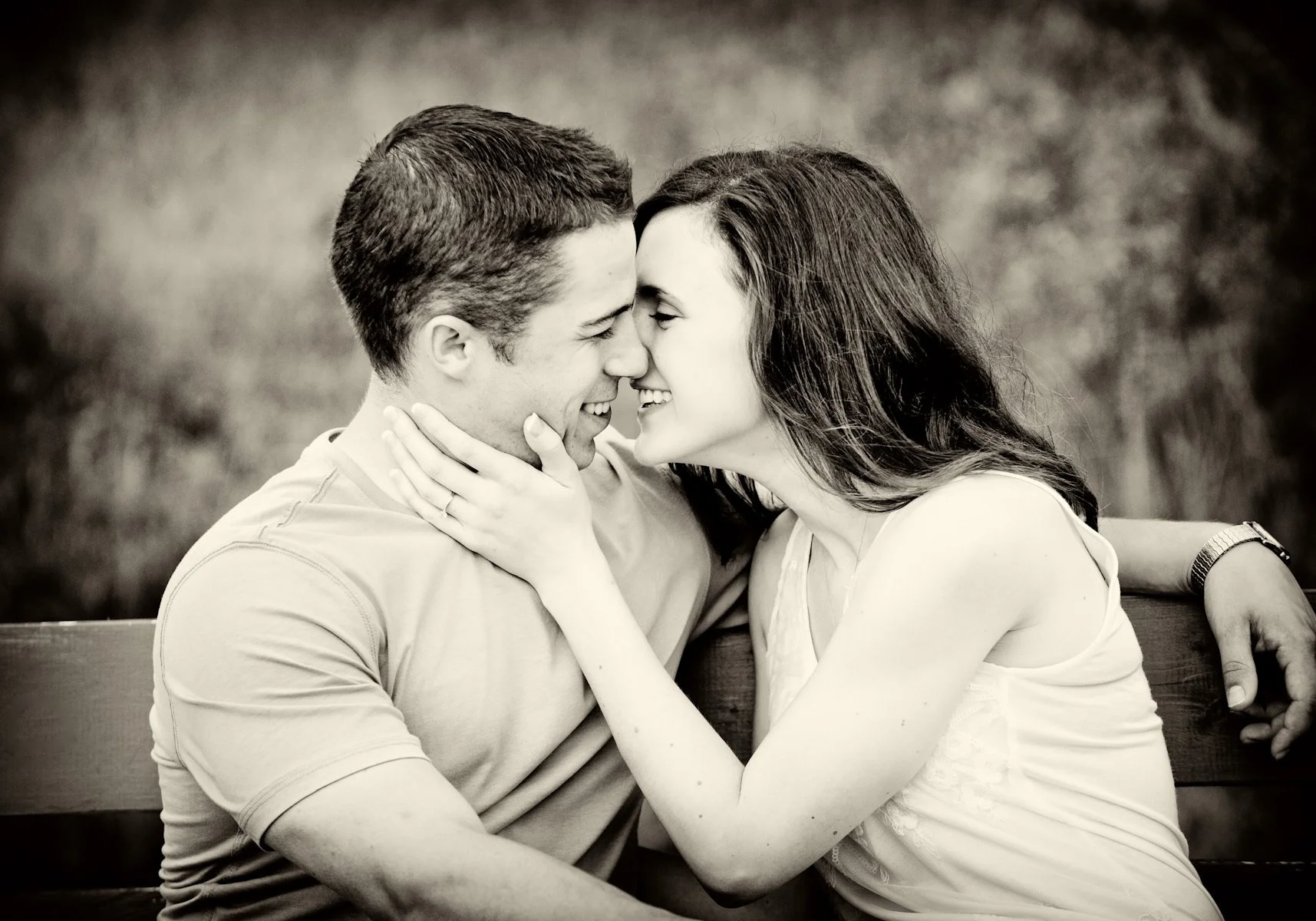 A black and white photo of a young man and woman sitting close together on a bench, touching foreheads and smiling, with the woman holding the man's face gently.