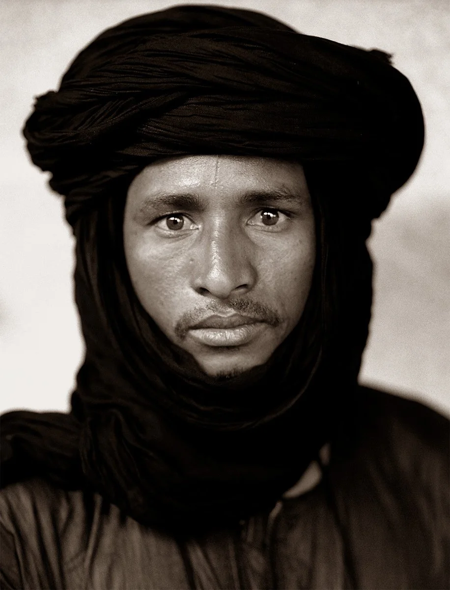 A black and white portrait of a man wearing a large, wrapped headscarf, looking directly at the camera.