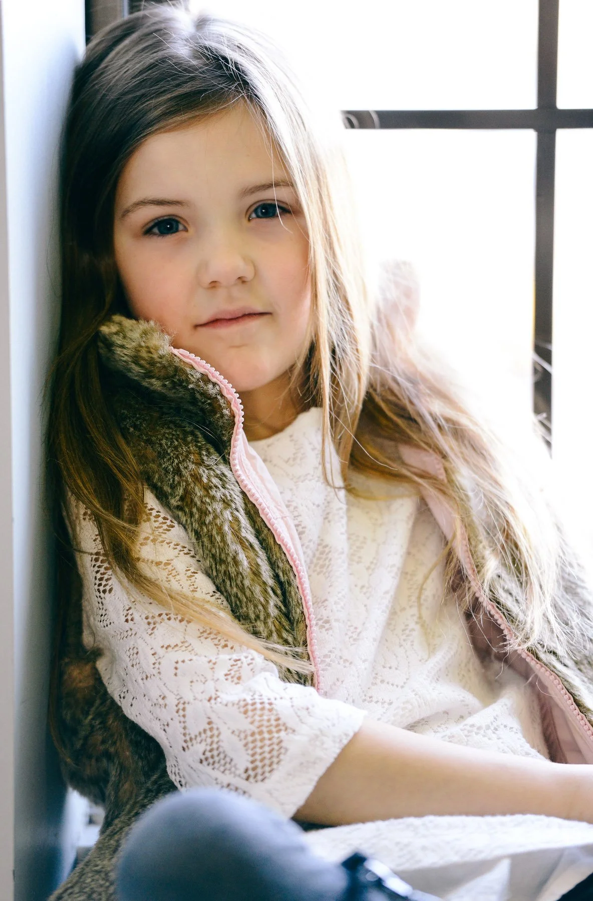 A young girl with long light brown hair and blue eyes sitting against a wall near a window. She's wearing a white lace dress and a jacket with a faux fur collar.
