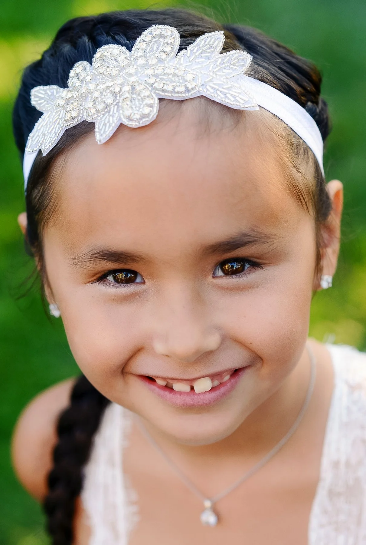 Close-up of a young girl with brown eyes, dark hair in a braid, wearing a white dress, a pearl necklace, and earrings, smiling outdoors with a green background.