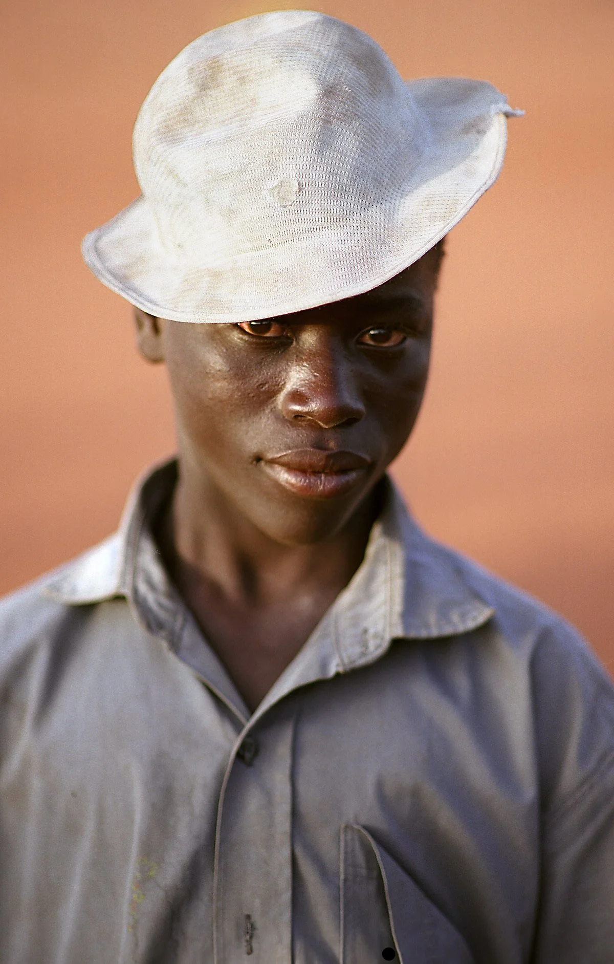 Portrait of a young man with dark skin wearing a white hat and gray shirt, set against a warm blurred background.