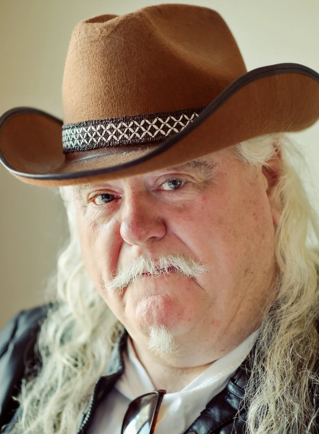 A close-up photo of a man with long white hair and a thick white mustache, wearing a brown cowboy hat with decorative band, a white shirt, and black leather jacket.