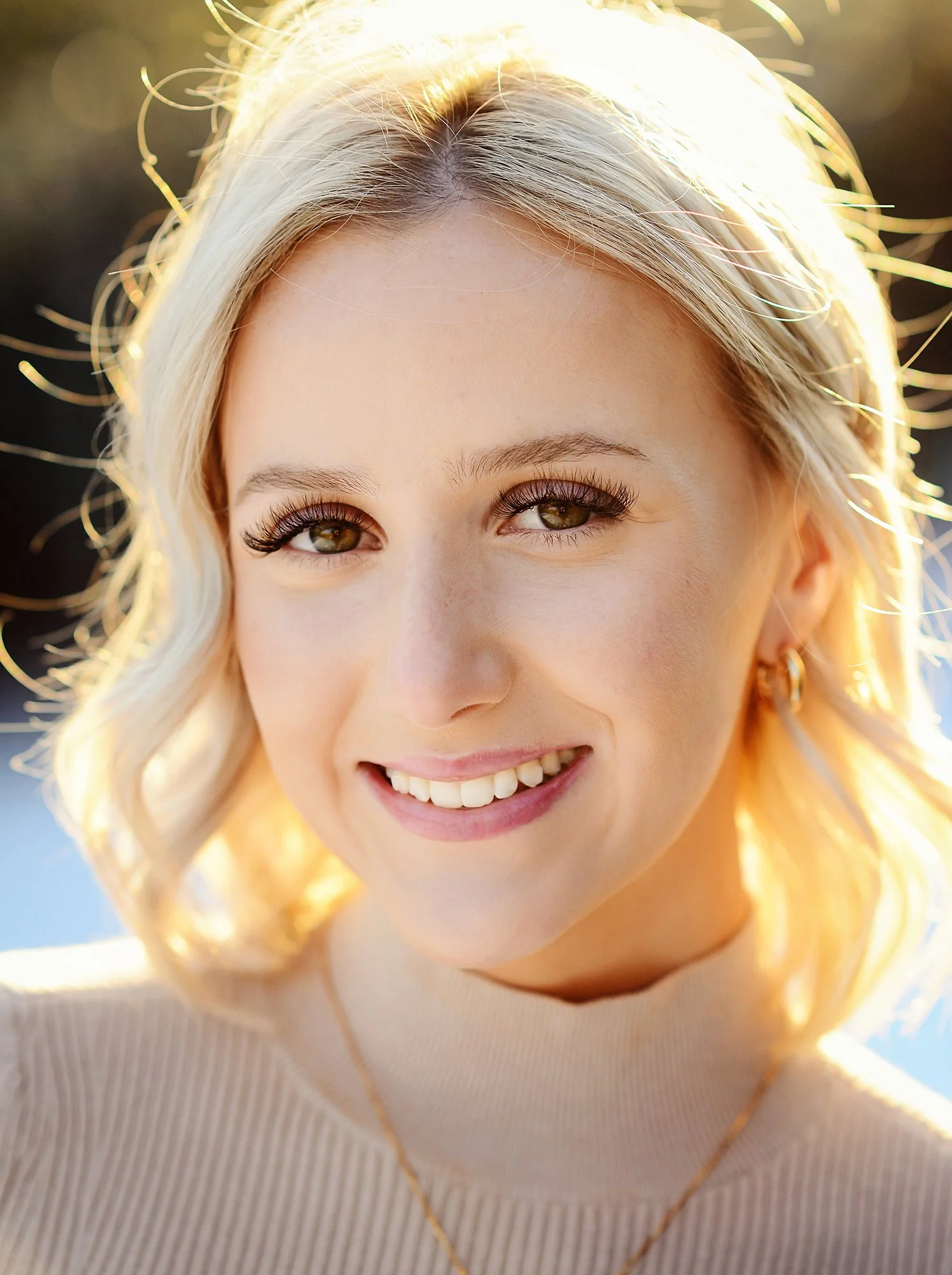Close-up of a smiling young woman with blonde hair, brown eyes, gold hoop earrings, wearing a beige top and gold necklace, backlit by sunlight.