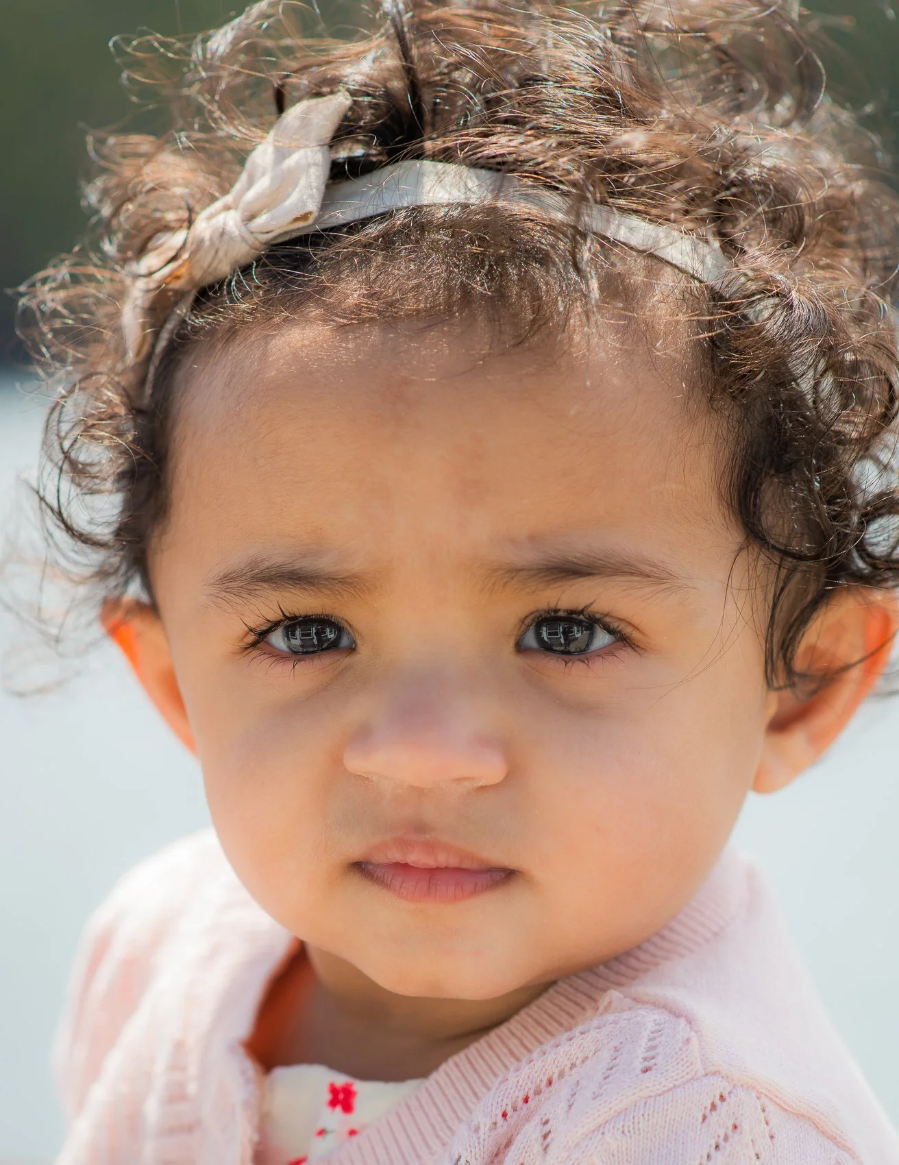 Close-up of a young girl with curly hair wearing a white headband and a light pink sweater, looking into the camera, with a soft, blurred background.