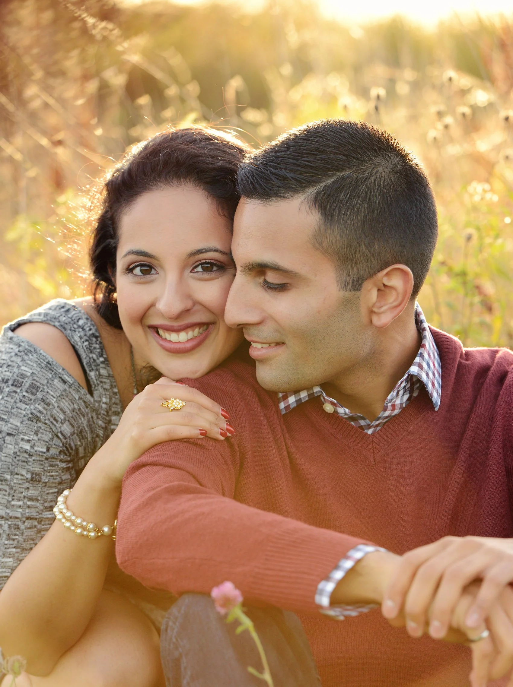 A smiling woman and a man are close together outdoors in a field during sunset, with sunny golden light in the background. The woman has dark hair, and the man has short dark hair and is wearing a red sweater over a checkered shirt.