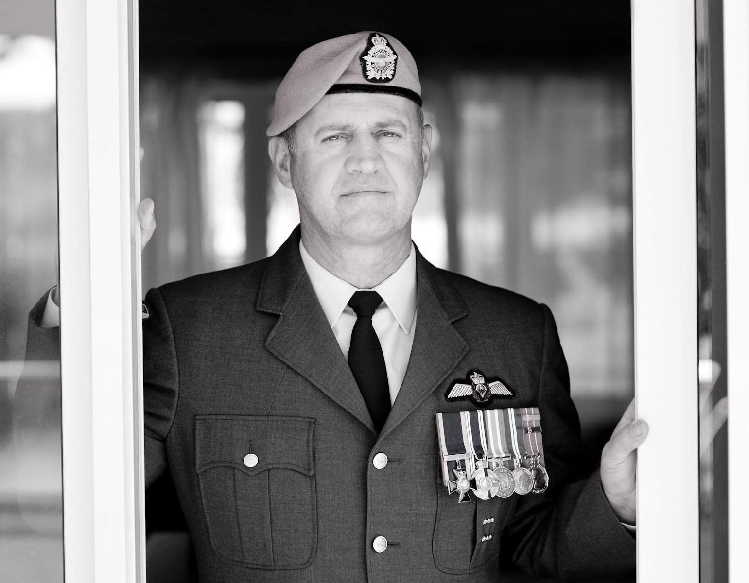Black and white photo of a man in a military uniform with medals, standing behind a glass door, wearing a beret, looking serious.