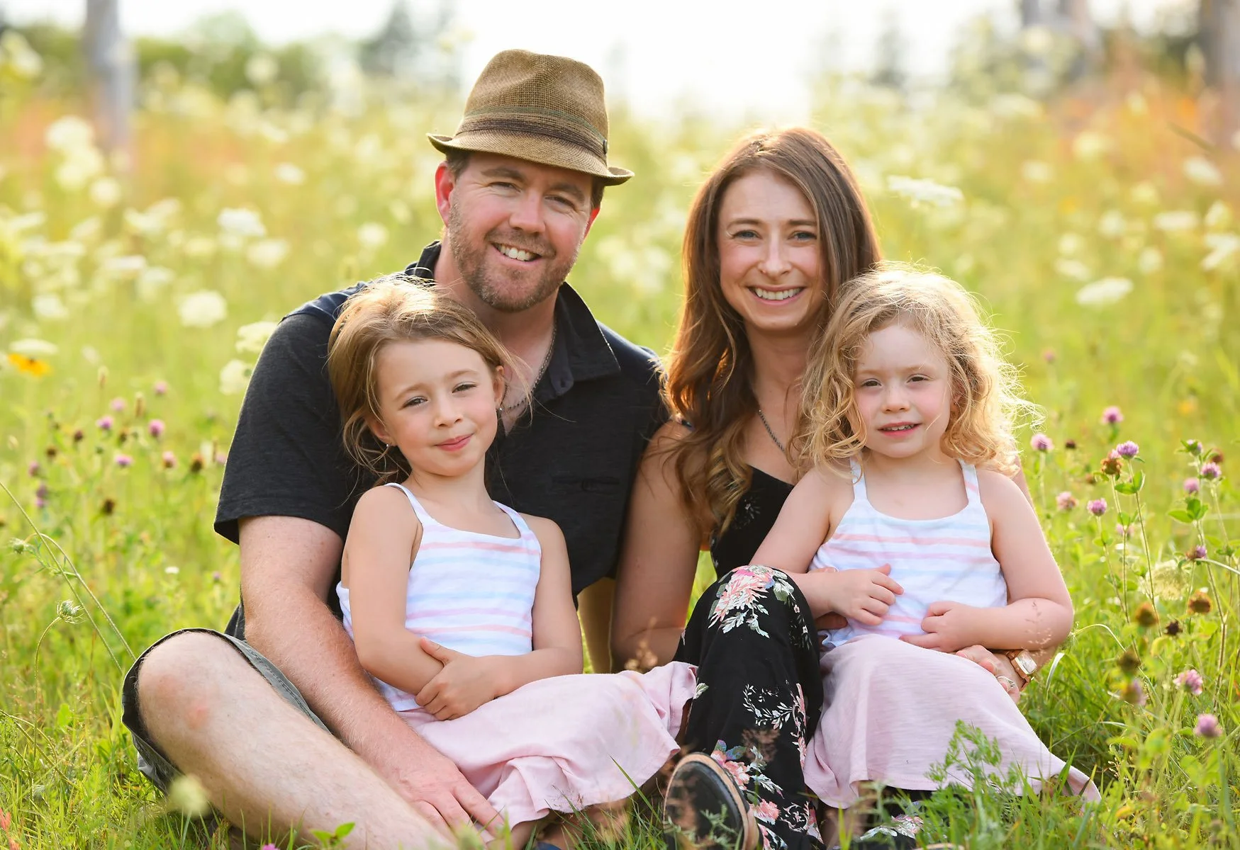 Family of four sitting together in a flower field during daytime, smiling at the camera.