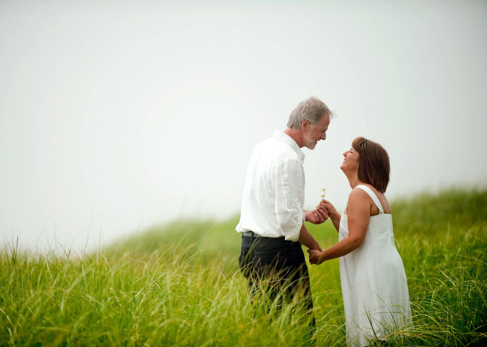An elderly man and woman sharing a romantic moment in a grassy field, with the man holding the woman's hand and the woman smiling at him, as he offers her a small flower.