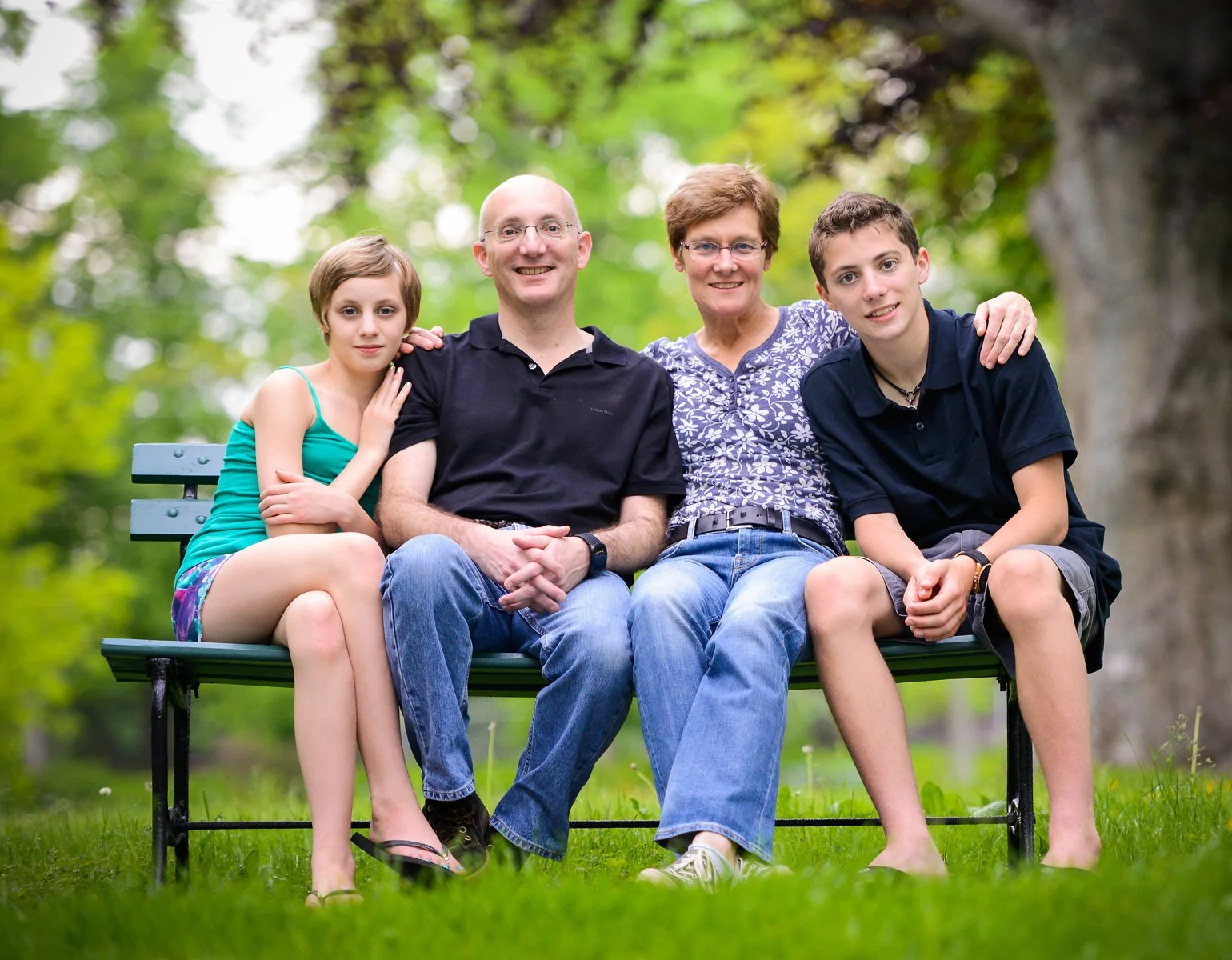 A family of four sitting on a park bench, smiling, with green trees in the background. The group includes a young girl, a middle-aged man and woman, and a teenage boy.
