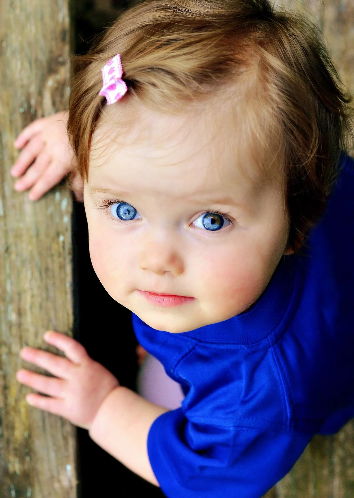 Close-up of a young girl with bright blue eyes, light brown hair with a pink hair clip, wearing a blue shirt, outdoors near a wooden surface, looking up at the camera.