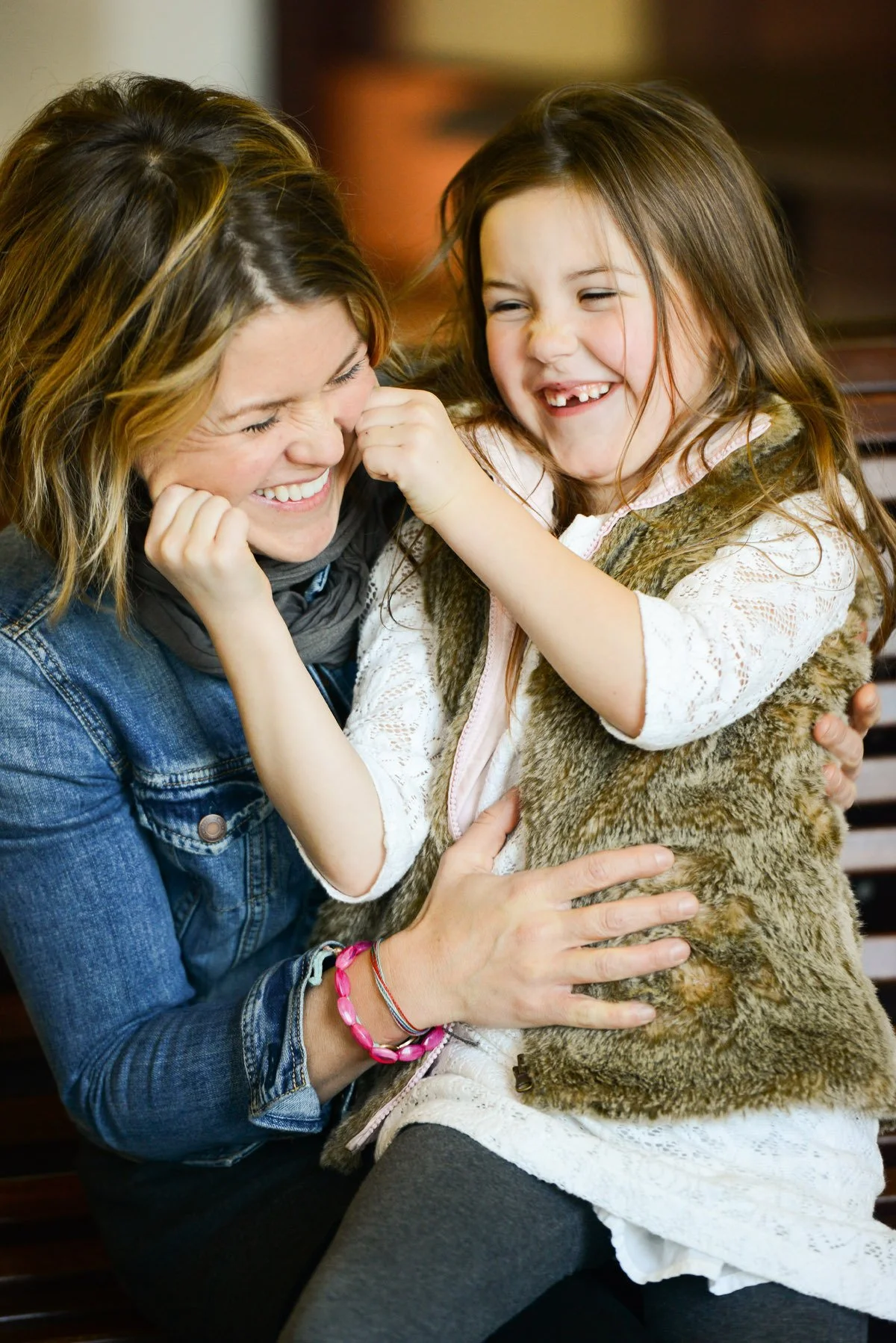 A young girl with missing teeth laughing and playingfully pinching an adult woman's cheeks, both smiling joyfully.