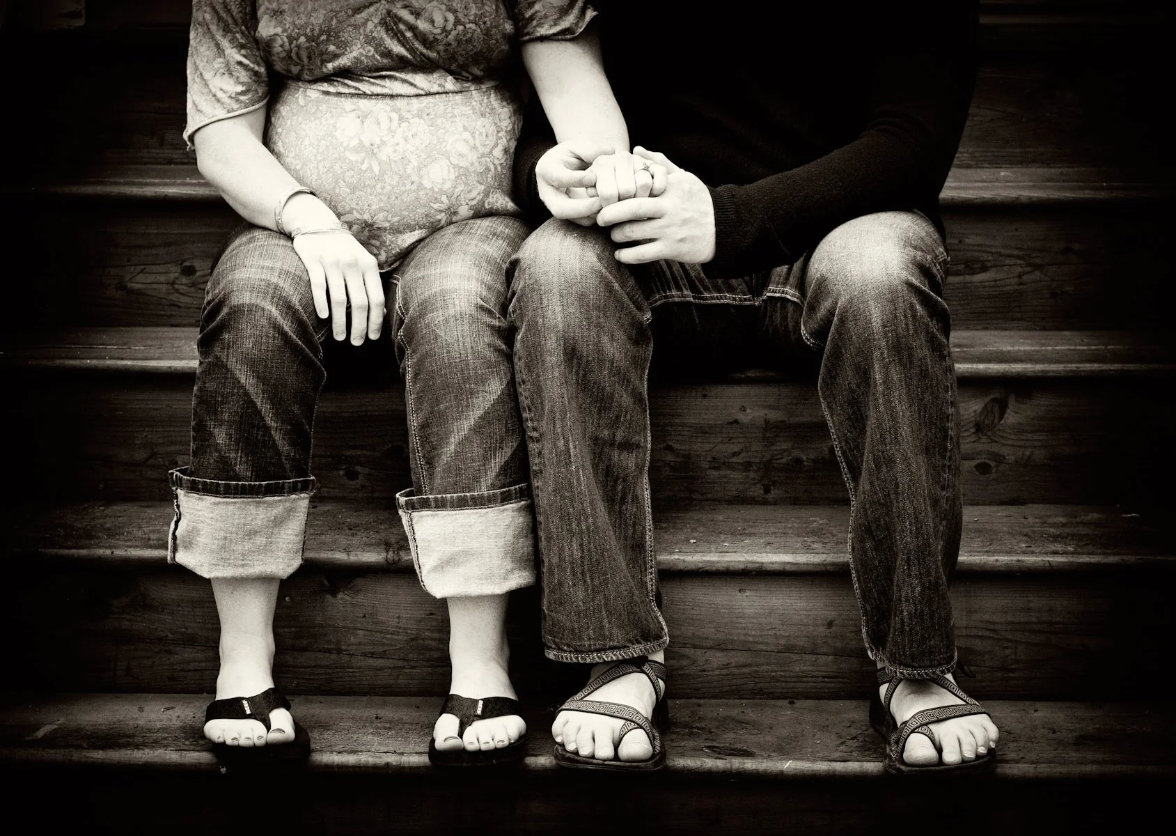 A black and white photo of a couple sitting on wooden steps, holding hands. The woman on the left is wearing rolled-up jeans and sandals, while the man on the right is dressed in jeans and sandals. Their hands are gently clasped with fingers intertwi
