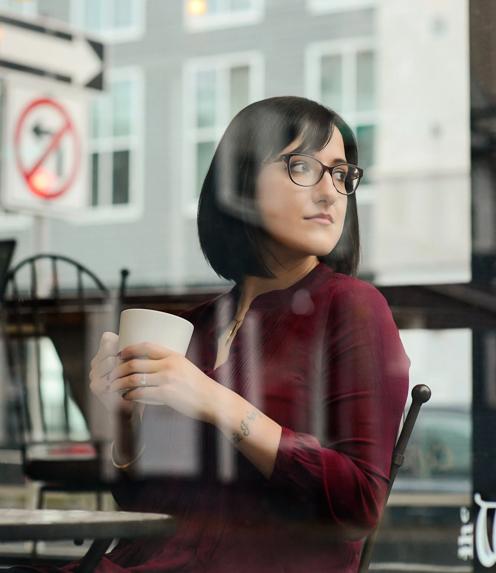 A woman with black hair and glasses sitting by a window, holding a white mug and looking outside.