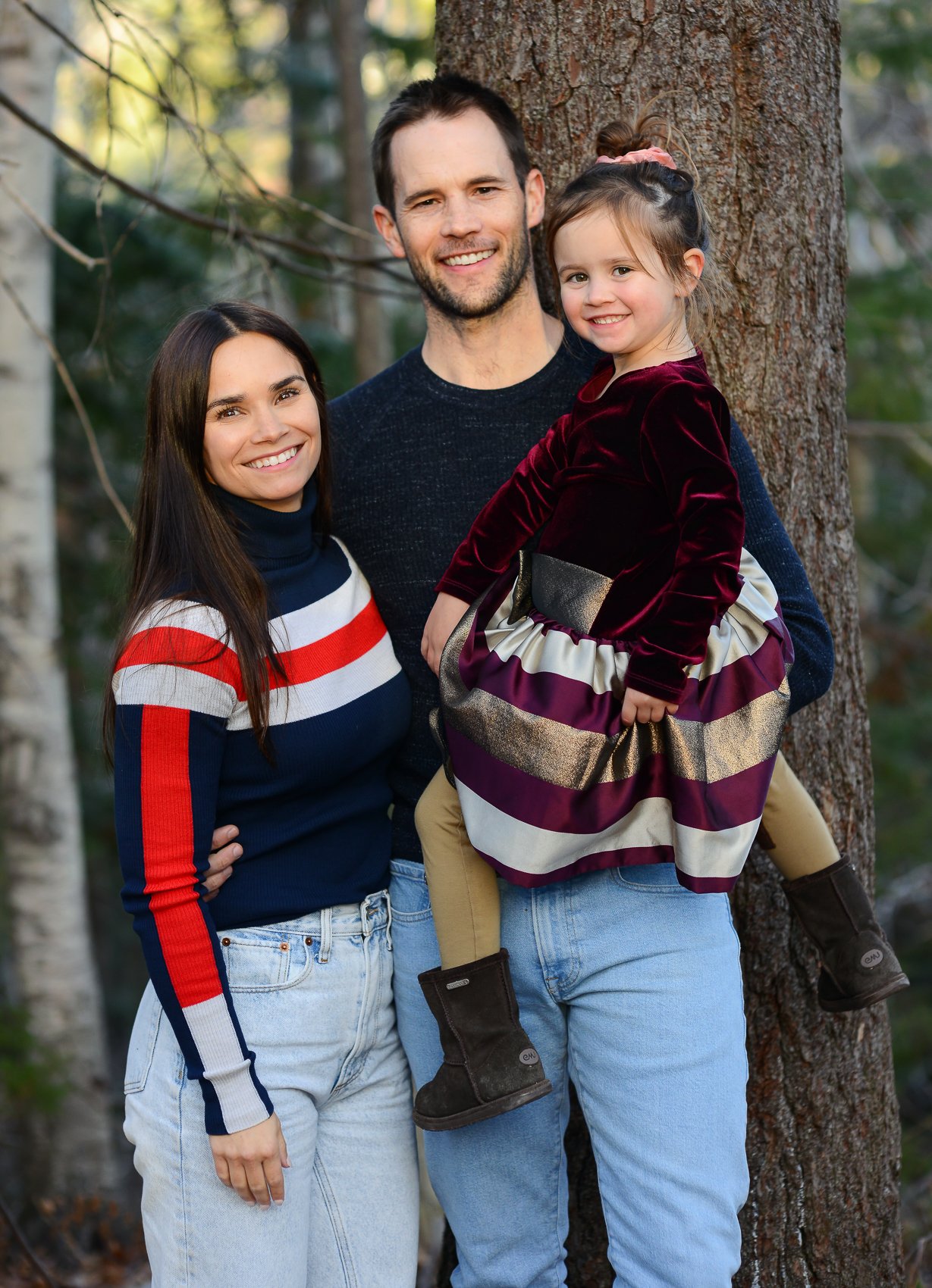 A family of three, with a woman, a man, and a young girl, posing outdoors in front of a tree in a forested area. They are smiling at the camera.
