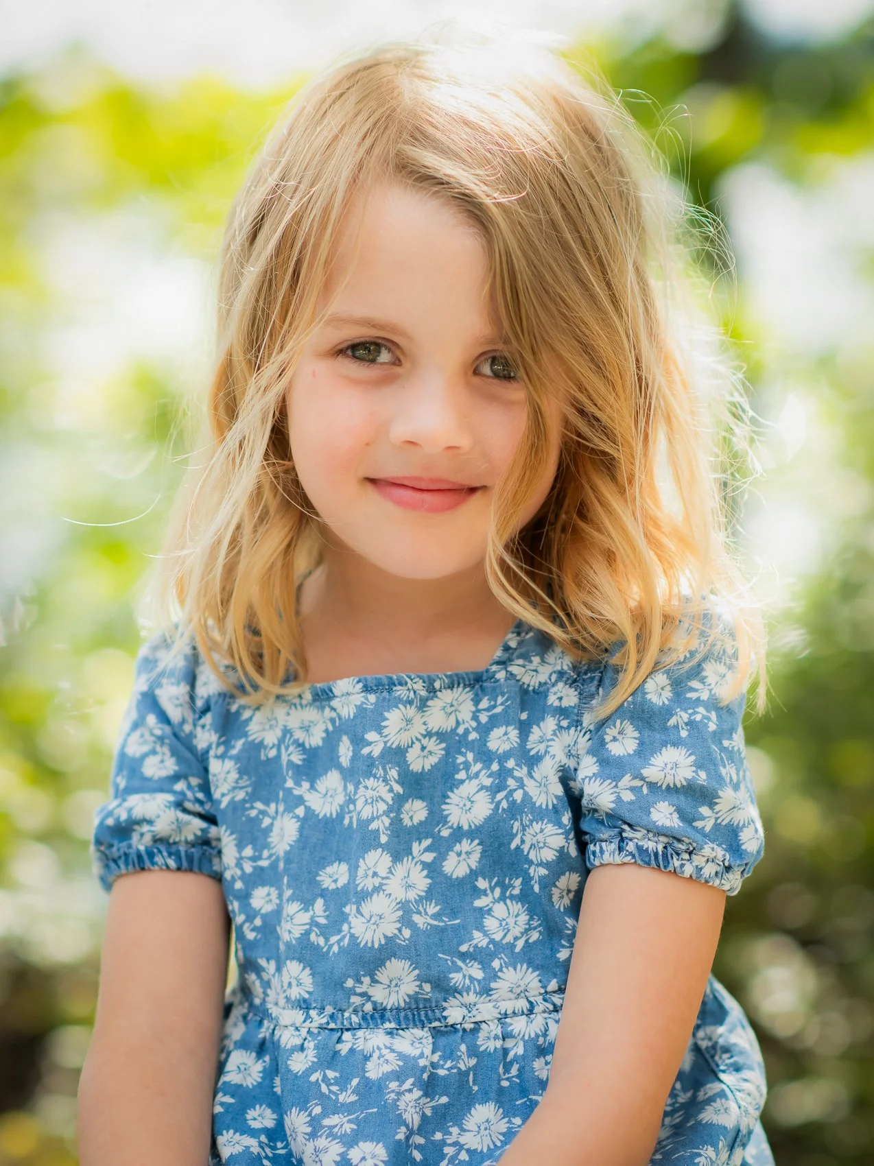 A young girl with blonde hair and blue eyes smiling outdoors, wearing a blue dress with white floral patterns.
