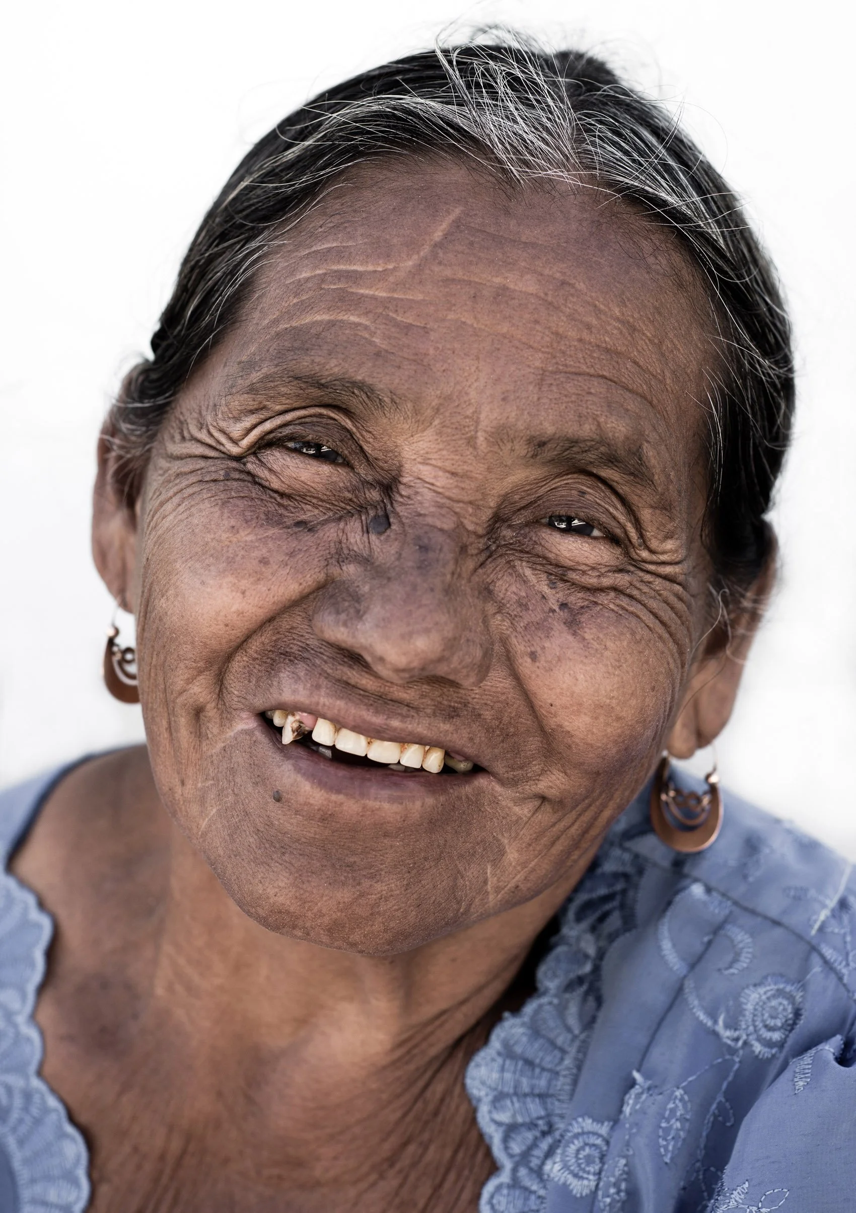 Close-up of a smiling elderly woman with gray hair, earrings, and a blue blouse, showing signs of aging and a missing tooth.
