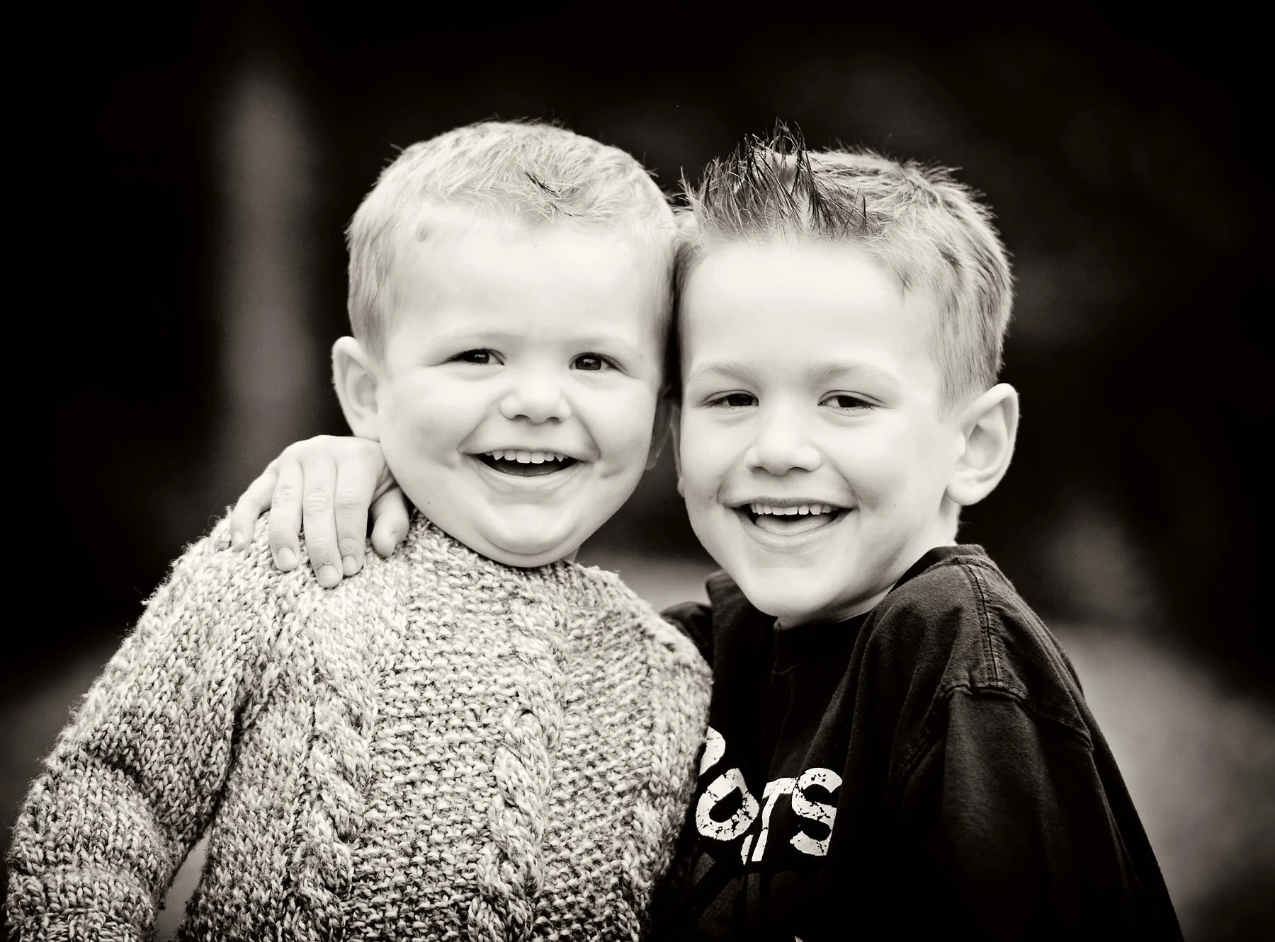 Two young boys smiling and hugging each other outdoors, one wearing a knitted sweater and the other in a black sweatshirt.