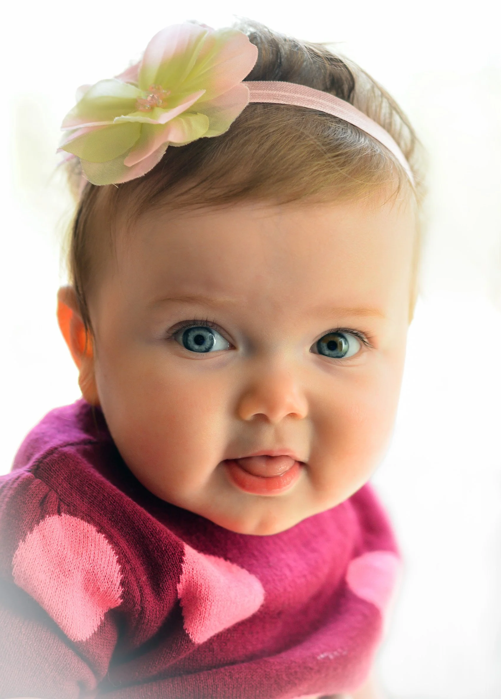 Close-up of a baby girl with blue eyes, wearing a pink headband with a large pink and yellow flower and a pink and purple sweater with heart patterns.